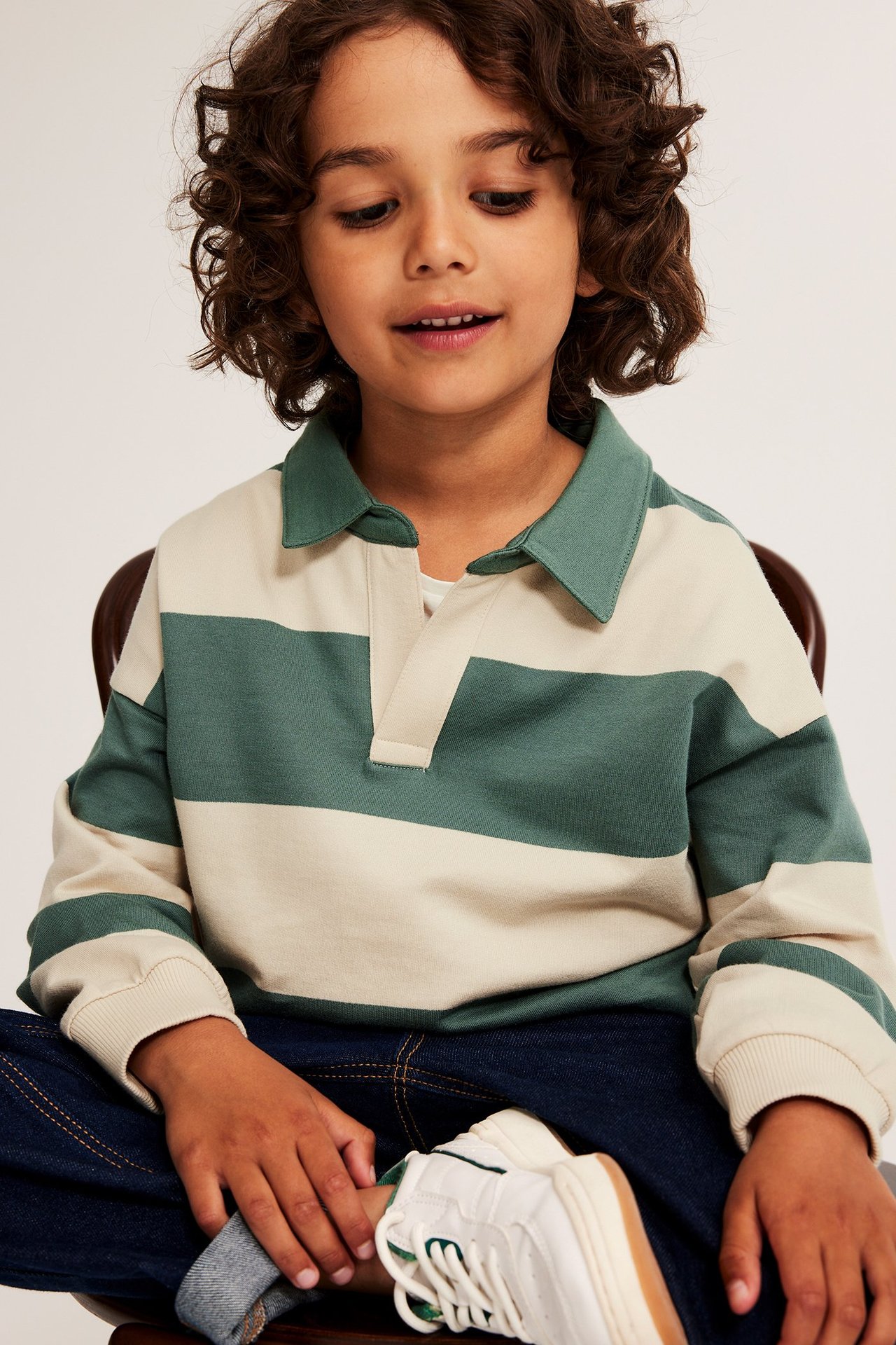 Frontbilde av et barn i stripete beige og grønn polo-genser, mørkeblå jeans og hvite sko.