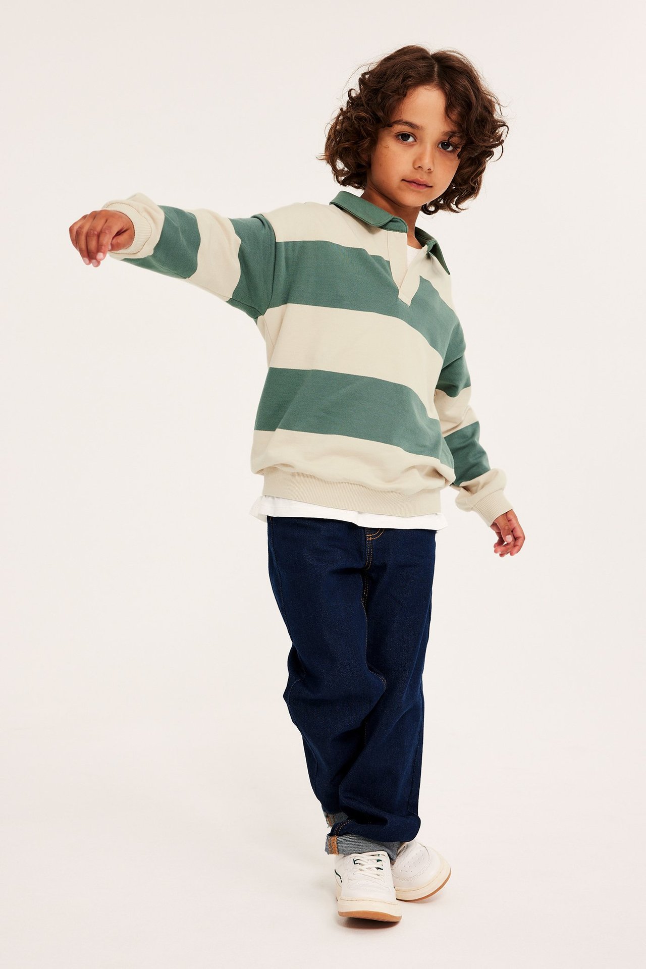 Widok z przodu dziecka w beżowo-zielonej bawełnianej bluzie polo w paski, ciemnych dżinsach i białych sneakersach.
