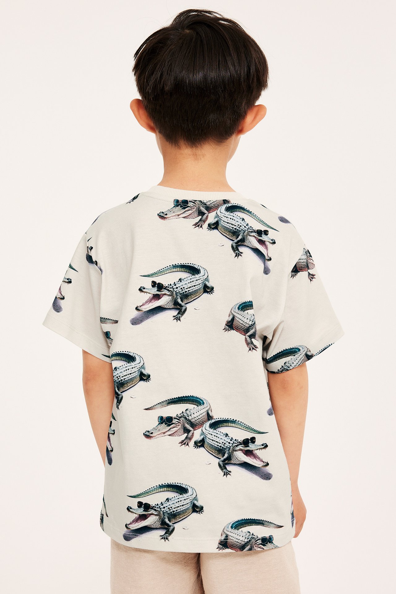 Bakifrån, ett barn i en ljusbeige T-shirt i bomull med heltäckande krokodilmönster.