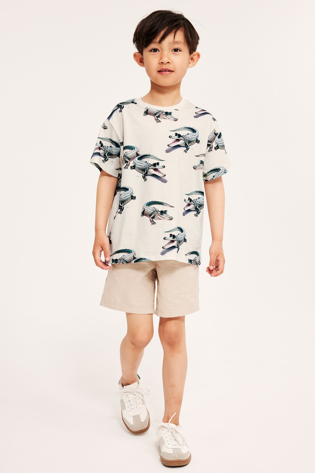 Framifrån: ett barn i ljusbeige bomulls-T-shirt med krokodilmönster, beiga shorts och vita sneakers.