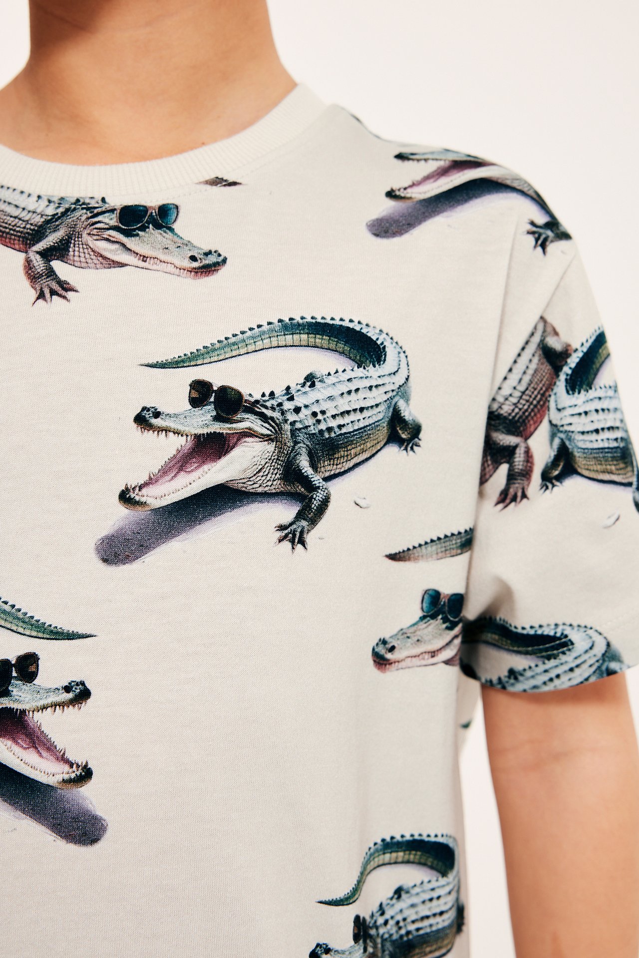 Närbild på ett barn i en ljusbeige bomulls-t-shirt med lekfullt alligatormönster och ribbad halsringning.