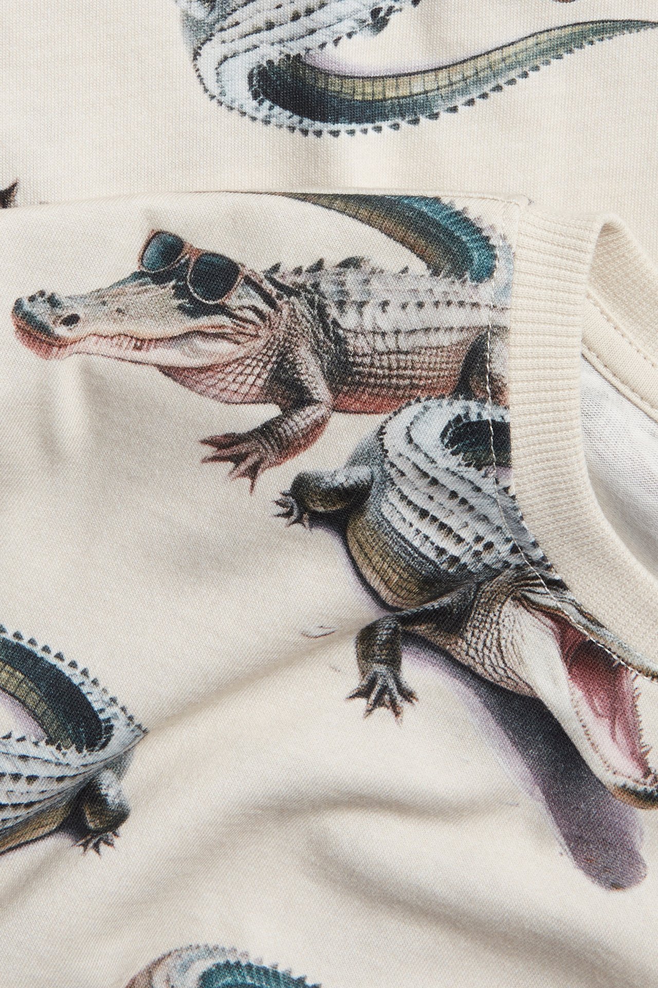 Närbild på en ljusbeige t-shirt i bomull med krokodilmönster, en krokodil bär solglasögon.