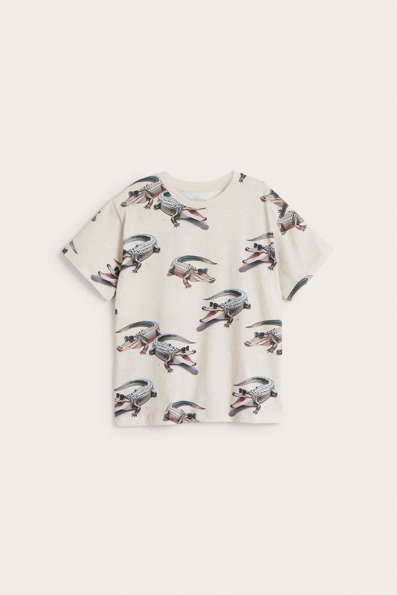 Frontvy av en ljusbeige t-shirt i bomull för barn med heltäckande krokodilmönster.