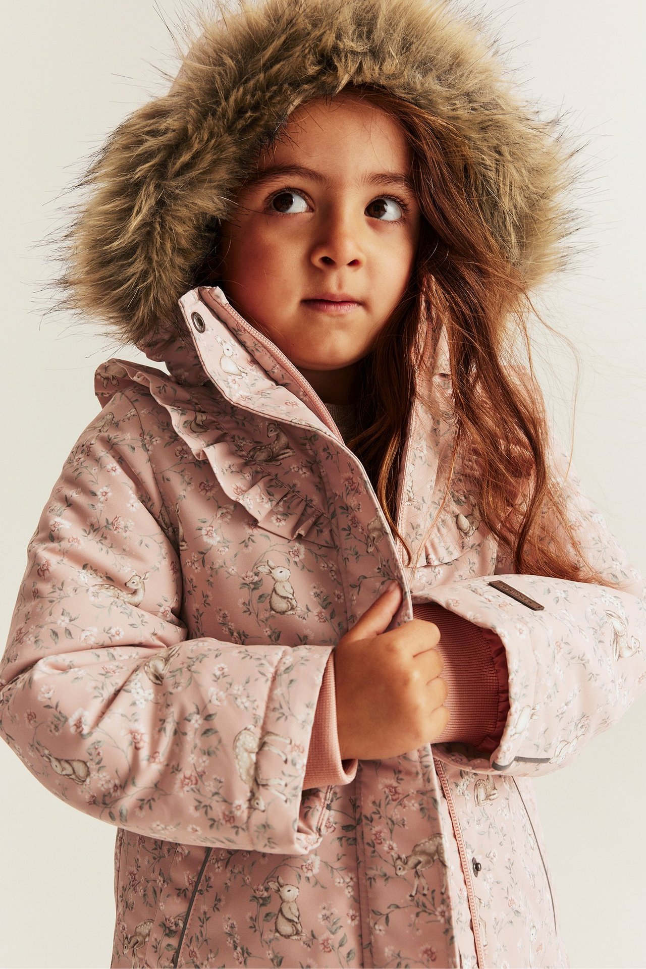 Widok z przodu: Dziecko w różowej, pikowanej kurtce w kwiaty z nadrukiem królików, futrzanym kapturem i falbanką.