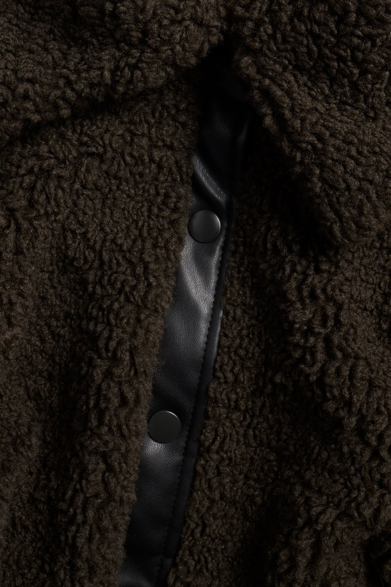 Zbliżenie ciemnobrązowej kurtki ze sztucznego futra z czarną listwą ze sztucznej skóry i zatrzaskami.