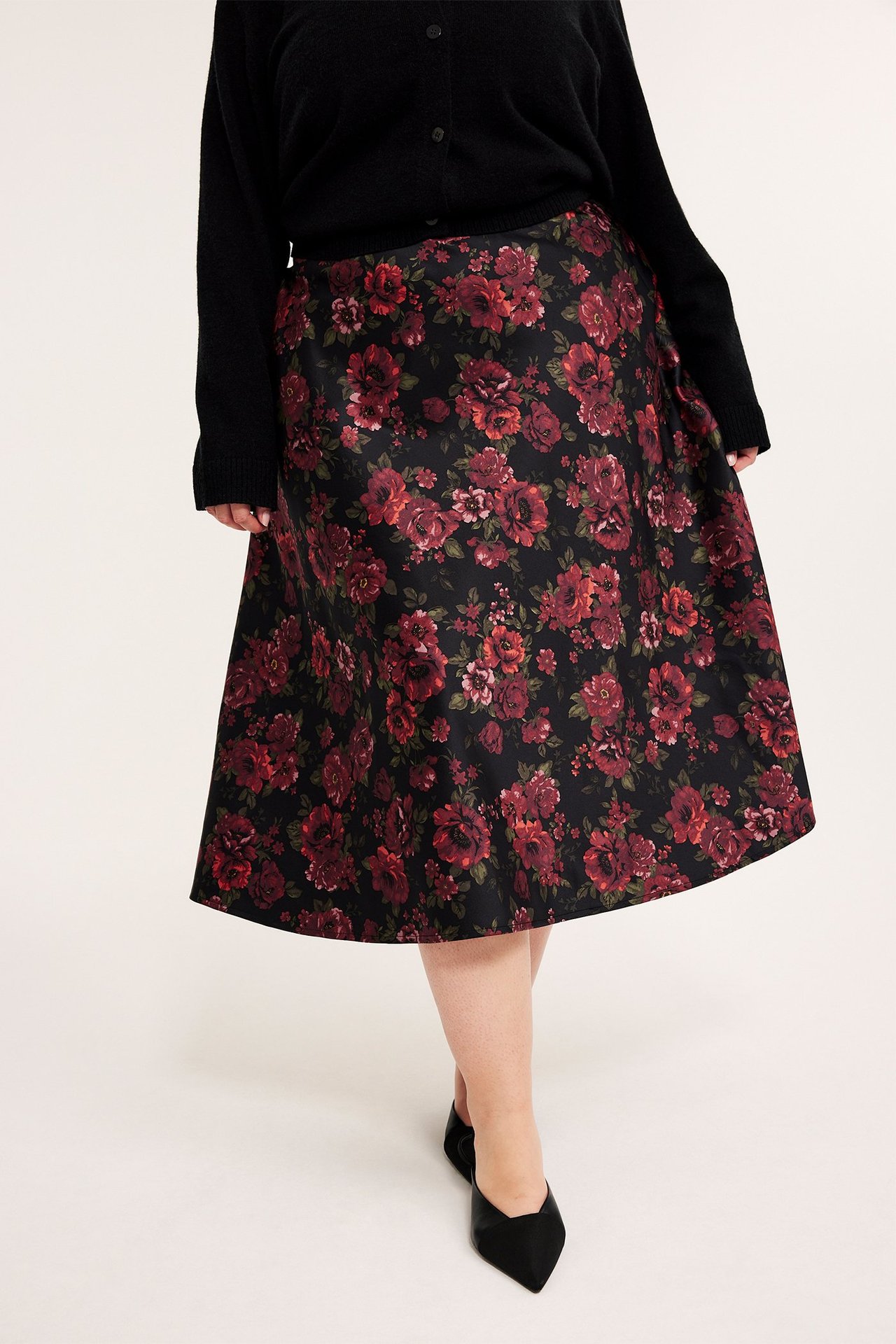 Framifrån, kvinna i svart utsvängd midikjol med rött blommönster, svart kofta och svarta platta skor.