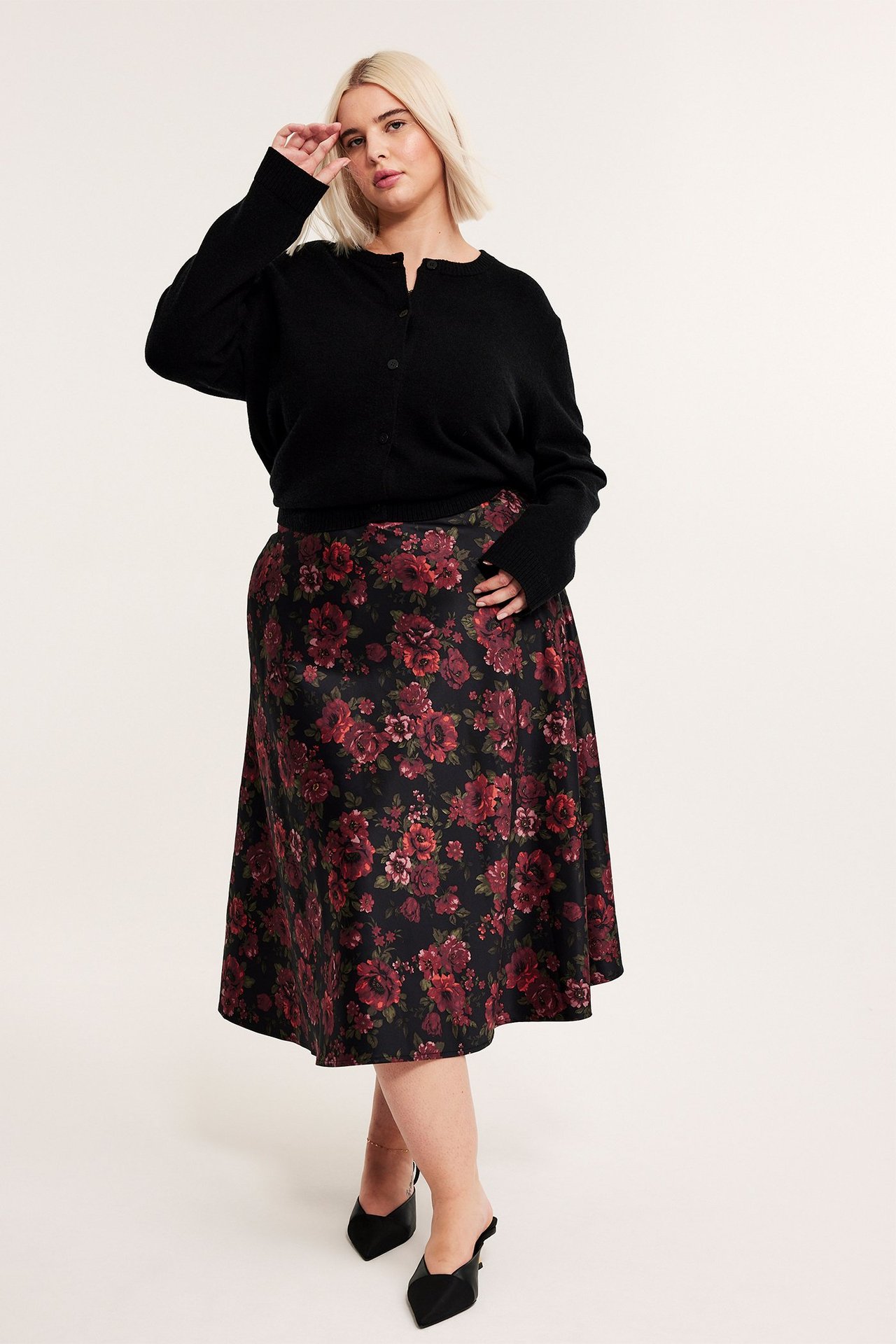 Widok z przodu: kobieta w czarnej kwiecistej rozkloszowanej spódnicy midi, czarnym kardiganie i czarnych klapkach.