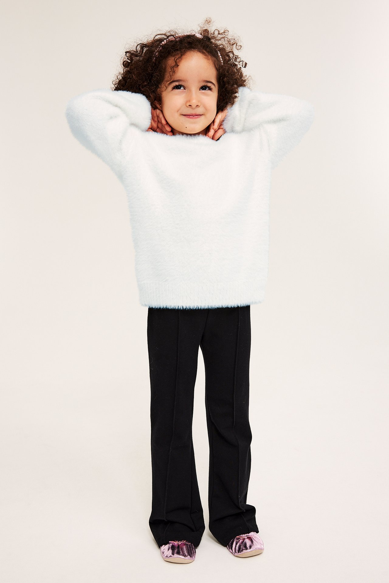 Frontvisning av et barn i en offwhite fluffy strikkegenser, svarte bukser og skinnende rosa sko.
