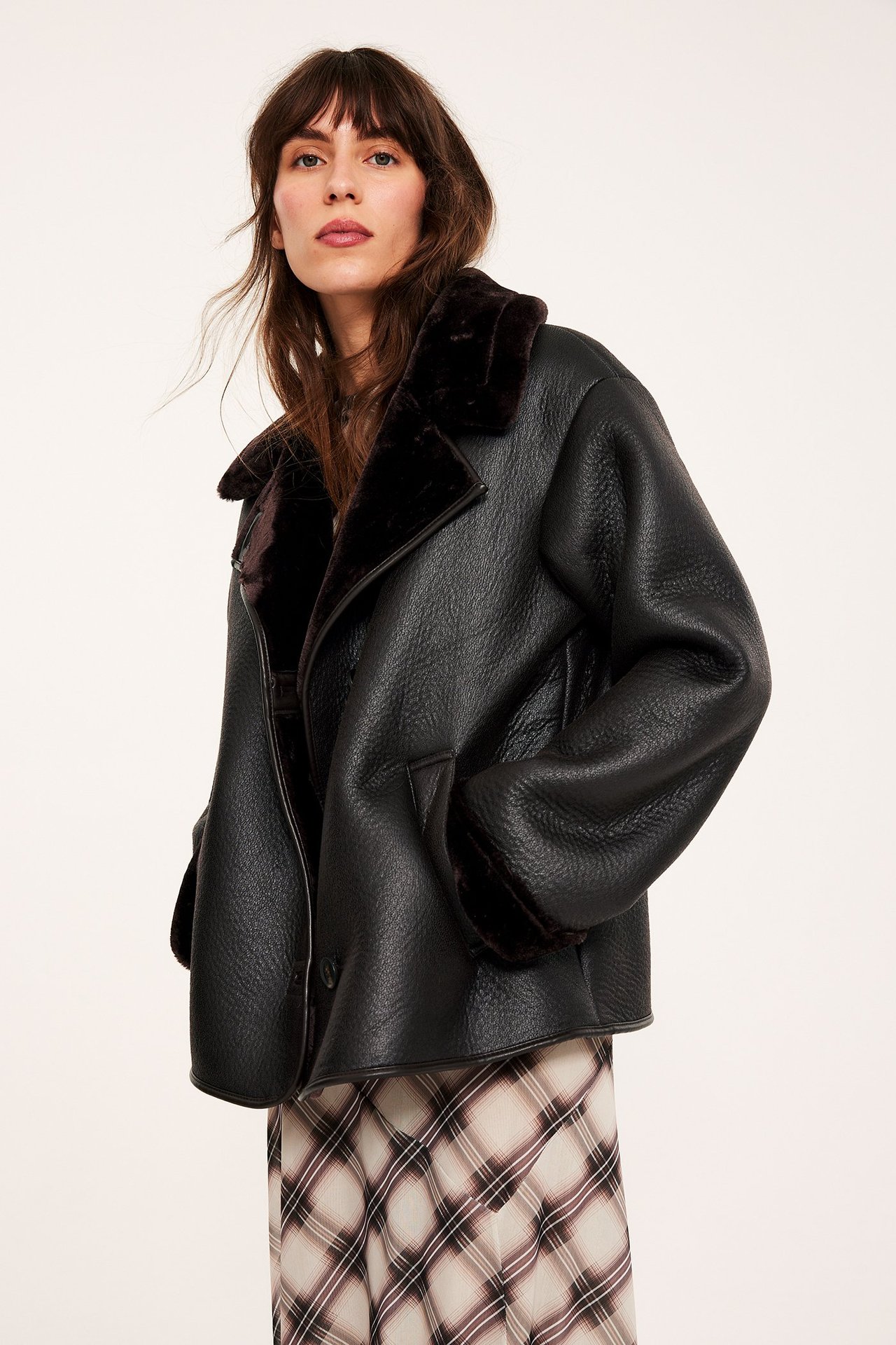 Edestä, nainen tummanruskeassa keinonahkaisessa shearling-takissa vaalean ruudullisen midihameen päällä.