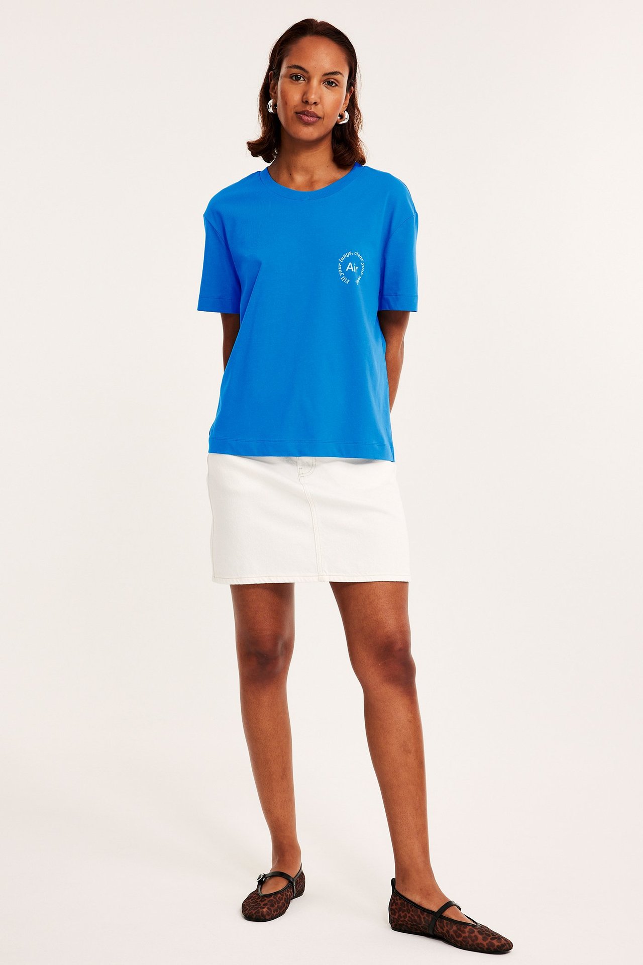 Forfra: kvinne i blå bomulls-T-skjorte med hvitt trykk, hvitt denim-miniskjørt og leopardmønstrede ballerinasko.