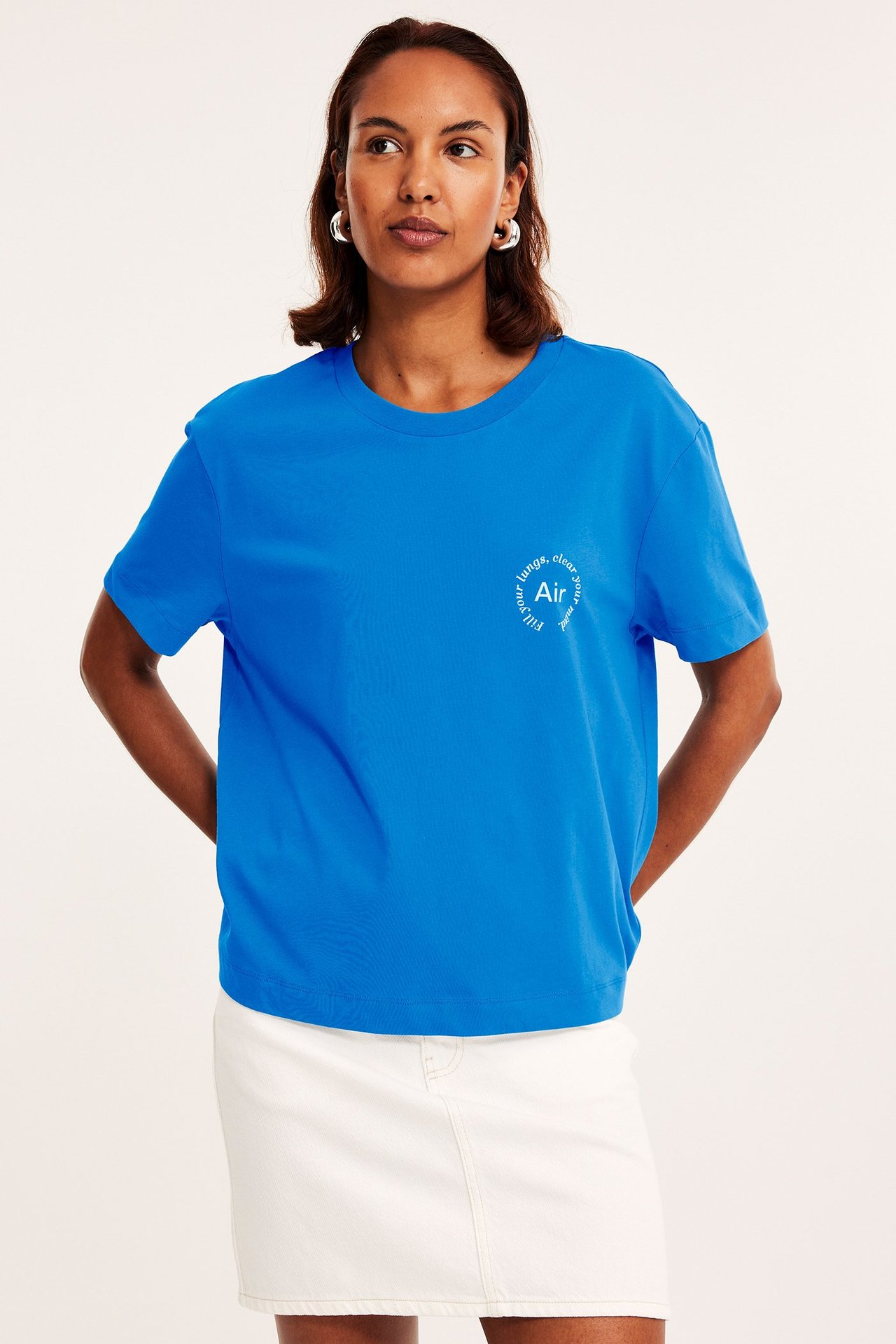 Kobieta w niebieskim bawełnianym T-shircie z białym nadrukiem, białej dżinsowej mini spódnicy i srebrnych kolczykach kołach.