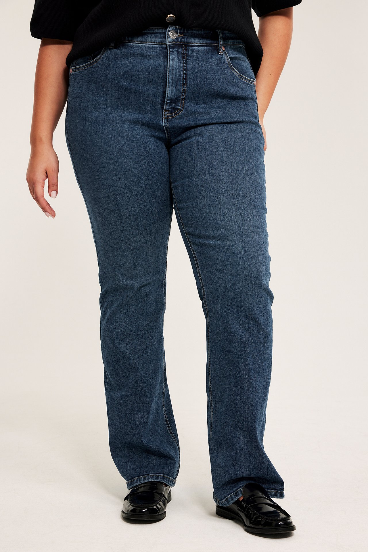 Edestä, nainen sinisissä korkeavyötäröisissä bootcut-farkuissa, musta toppi ja mustat loaferit.