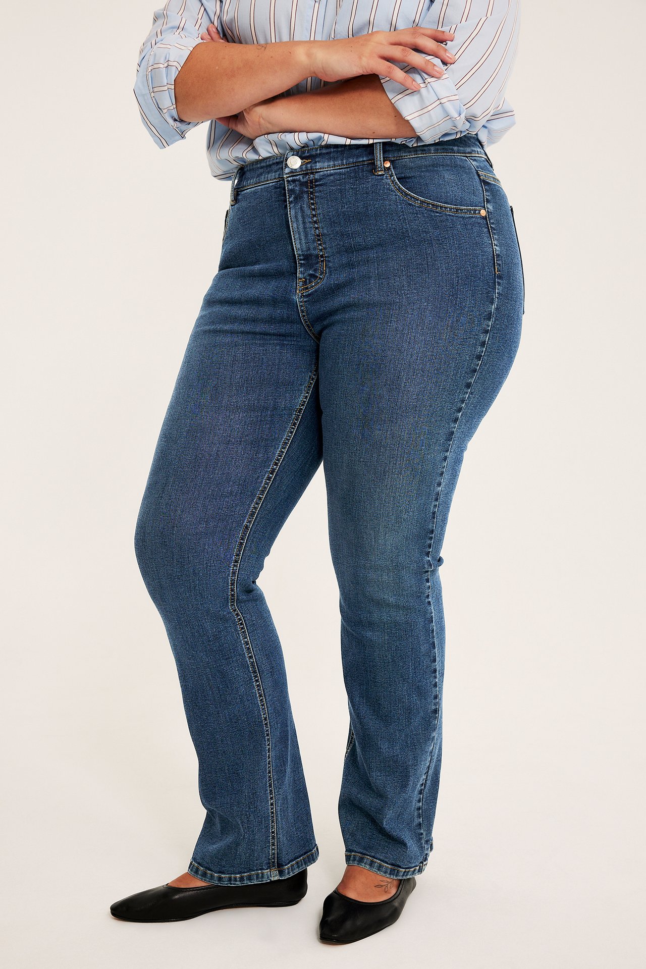 Sidevisning av en kvinne i blå bootcut-jeans, en lyseblå stripete skjorte og svarte flate sko.