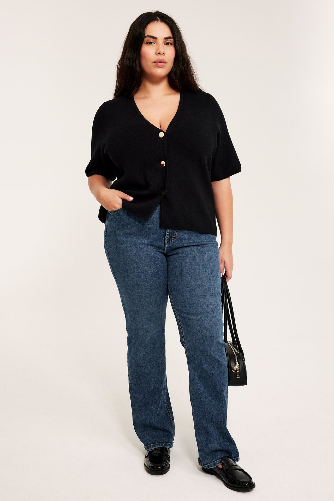 Edestä: Nainen sinisissä korkeavyötäröisissä bootcut-farkuissa, musta lyhythihainen napitettu toppi, mustat loaferit, musta käsilaukku kädessä.
