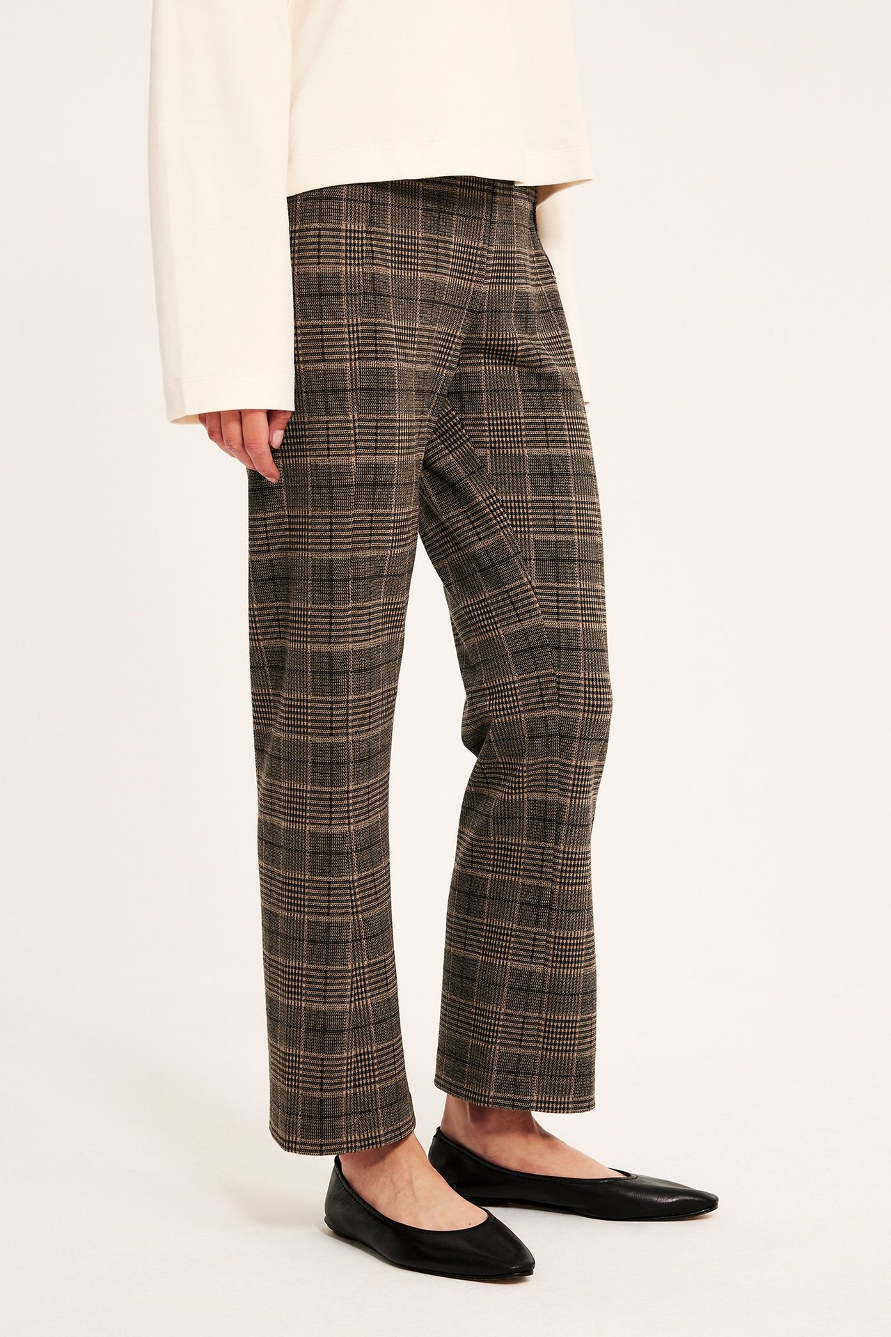 Sivukuva naisesta yllään ruskeat ruudulliset pull-on kickflare-housut, kermanvärinen toppi ja mustat ballerinat.