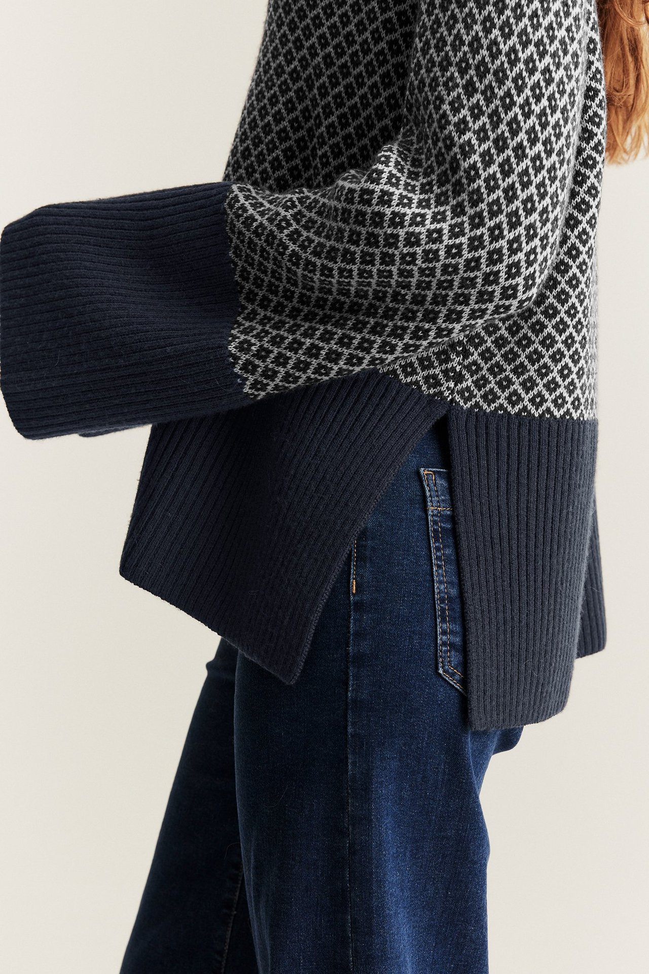 Sidovy av kvinna i mörkblå jacquardmönstrad ulltröja med vida ärmar och mörkblå jeans.