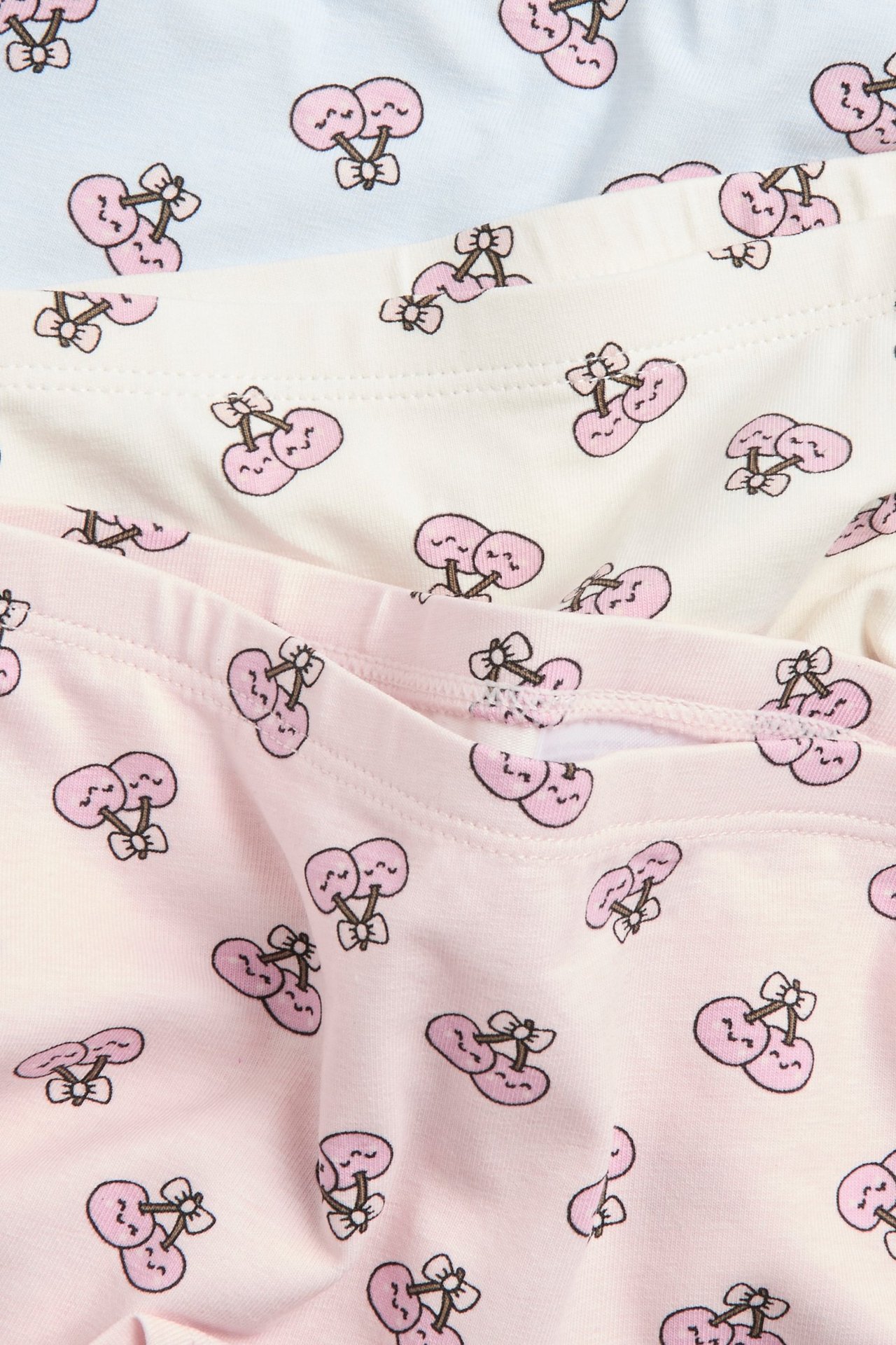 Zbliżenie na trzy pary dziecięcych bokserek: jasnoniebieskie, jasnobeżowe i jasnoróżowe, z różowymi wiśniami.