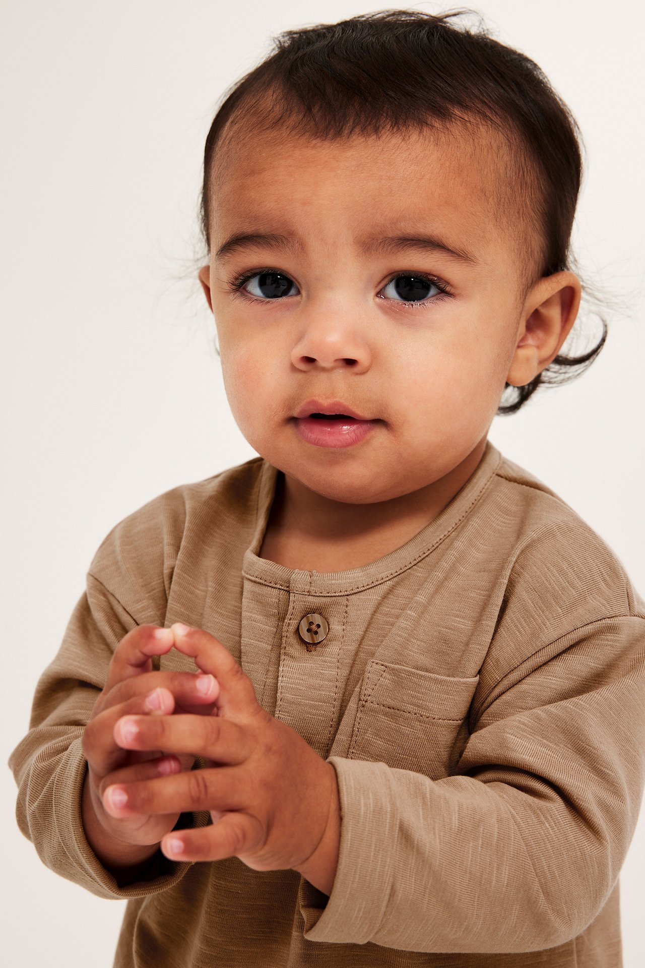 Närbild framifrån på en baby i en brun långärmad jerseyt-shirt med knappar och bröstficka.