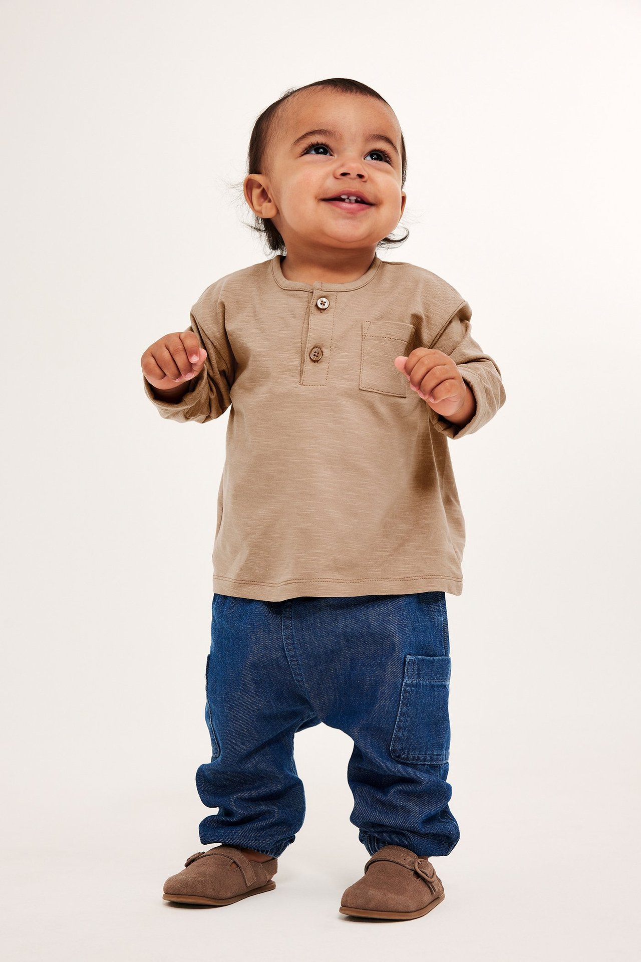 Widok z przodu: niemowlę w brązowej koszulce z długim rękawem, niebieskich dżinsach i brązowych butach.