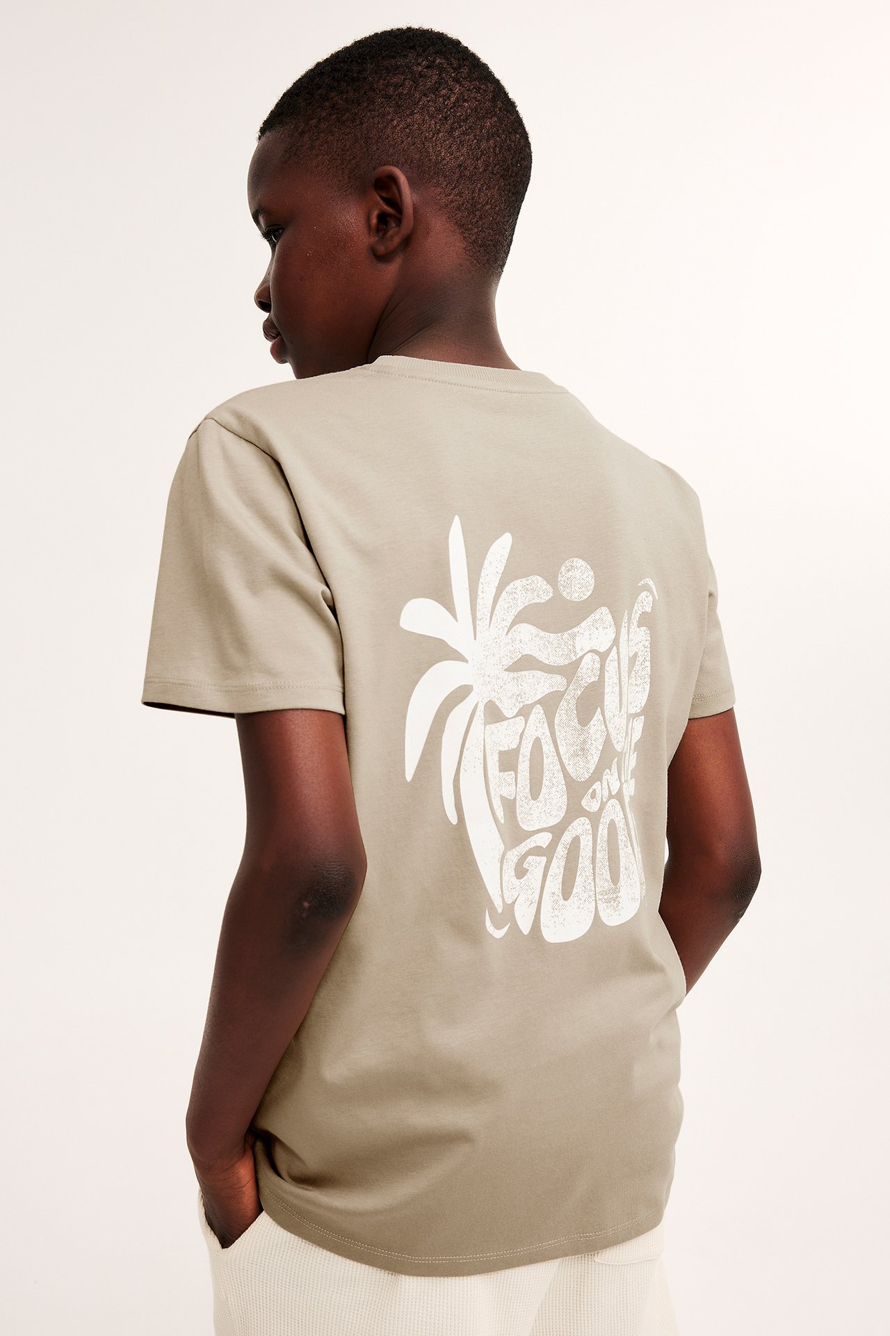 Bakifrån: ett barn i en beige bomulls-T-shirt med stort vitt grafiskt tryck på ryggen.