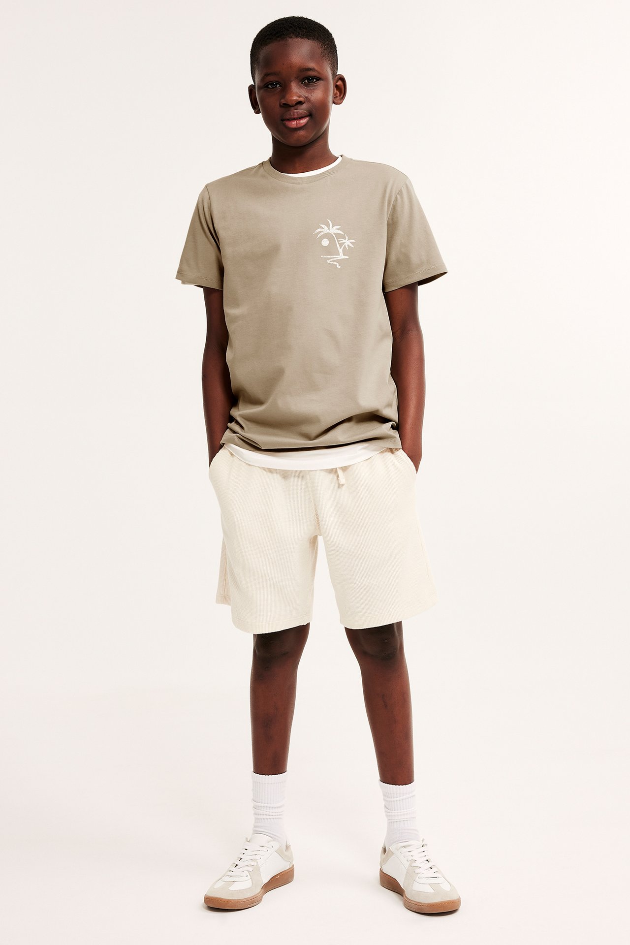 Framifrån: beige bomullst-shirt med palmtryck, cremefärgade shorts, vita strumpor och sneakers.