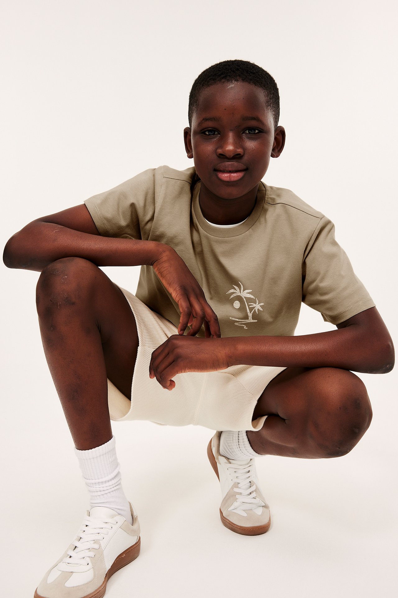 Framifrån: Ett barn i beige bomulls-T-shirt med palmträdtryck, ljusa ribbade shorts och sneakers.