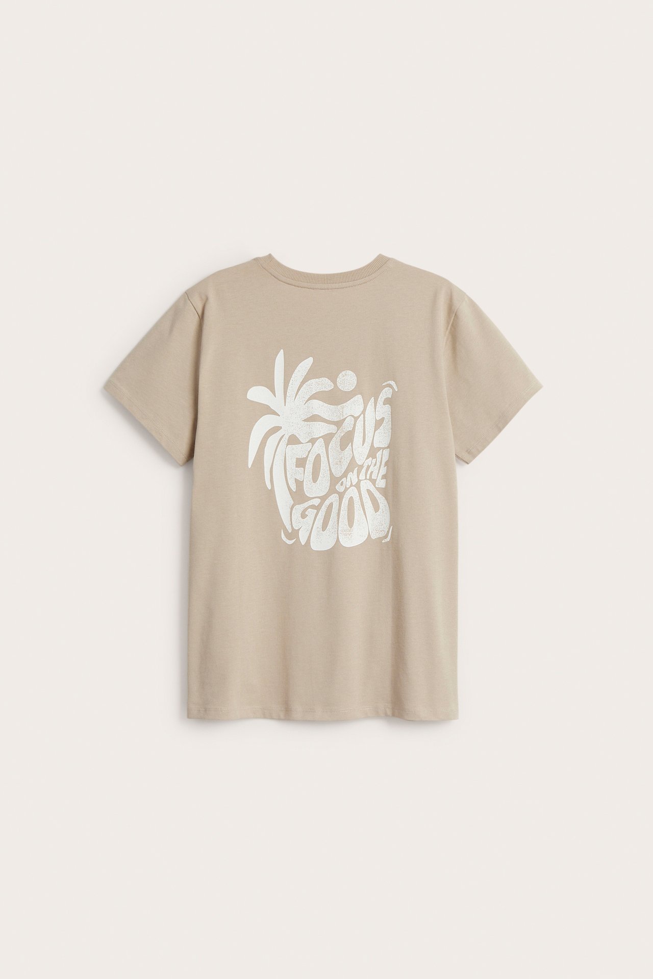 Bakifrån av en beige bomulls-t-shirt med stort vitt "Focus on the Good" palmträdtryck.