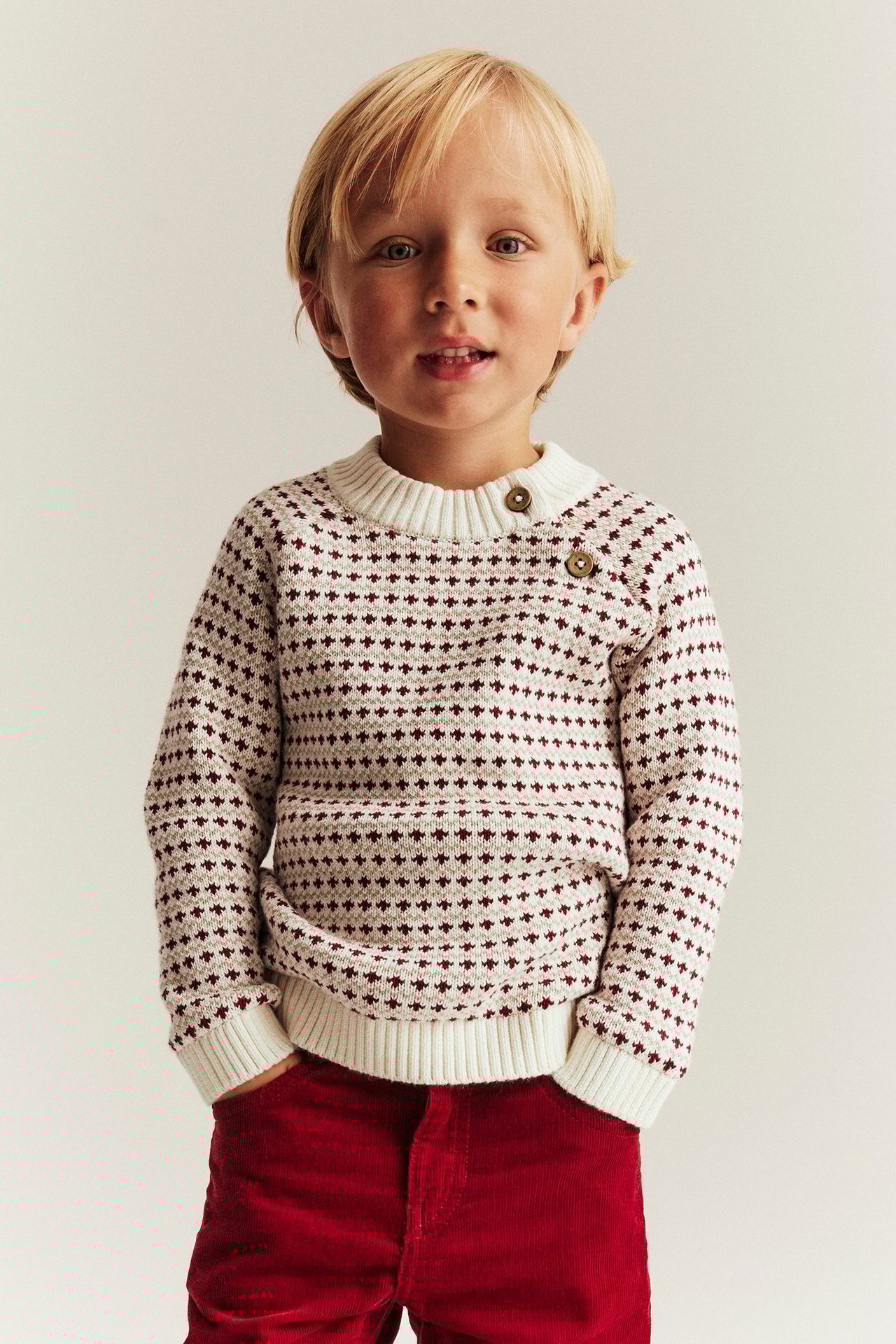 Poika yllä offwhite-puuvillaneule, punainen jacquard-kuvio, napit, punaiset farkut.