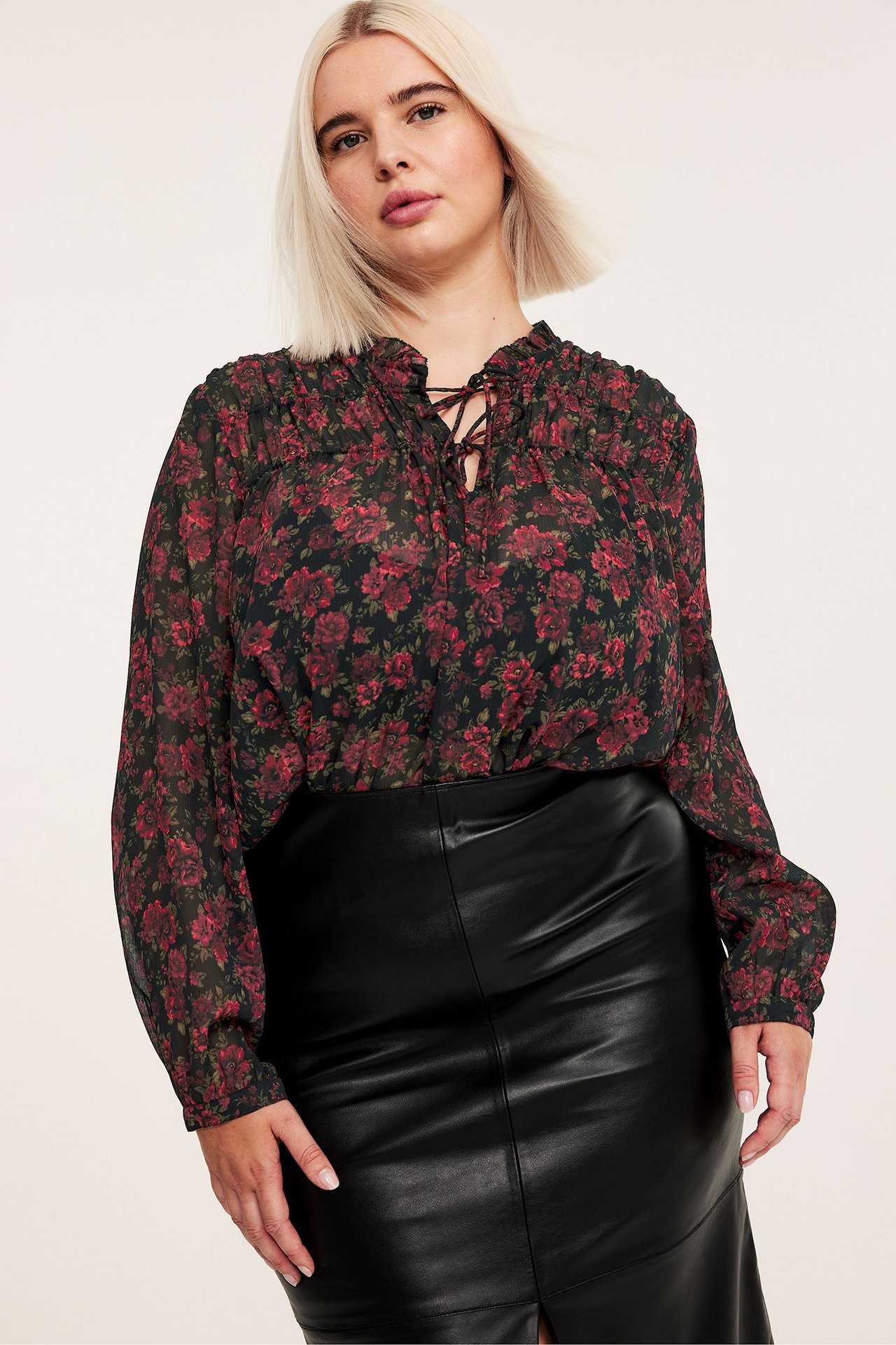 Widok z przodu: Kobieta w czarnej wzorzystej bluzce z długim rękawem i sznurowaniem, oraz czarnej spódnicy z ekoskóry.