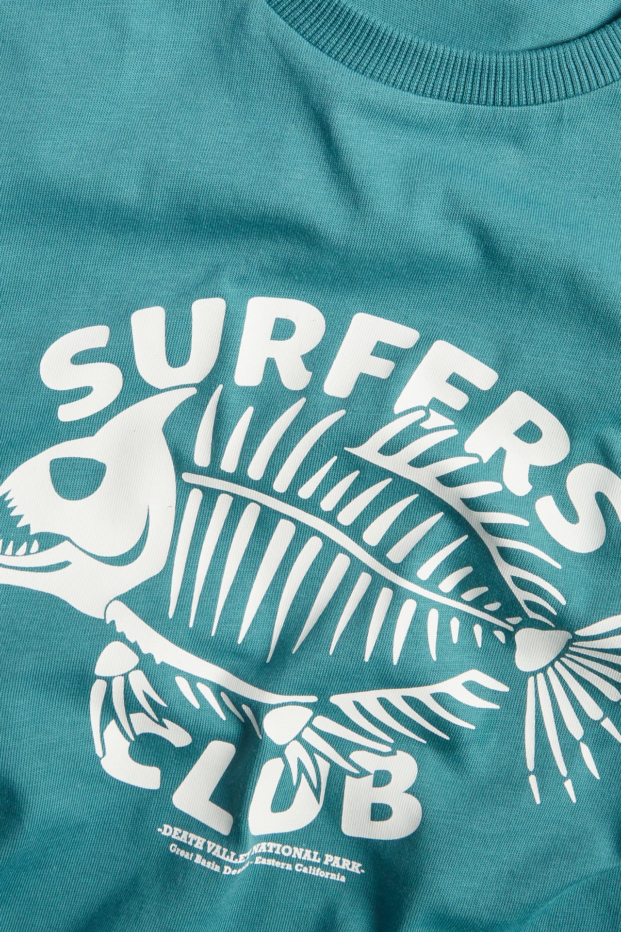 Zbliżenie z przodu zielonej bawełnianej koszulki z białym nadrukiem "SURFERS CLUB" i szkieletem ryby.