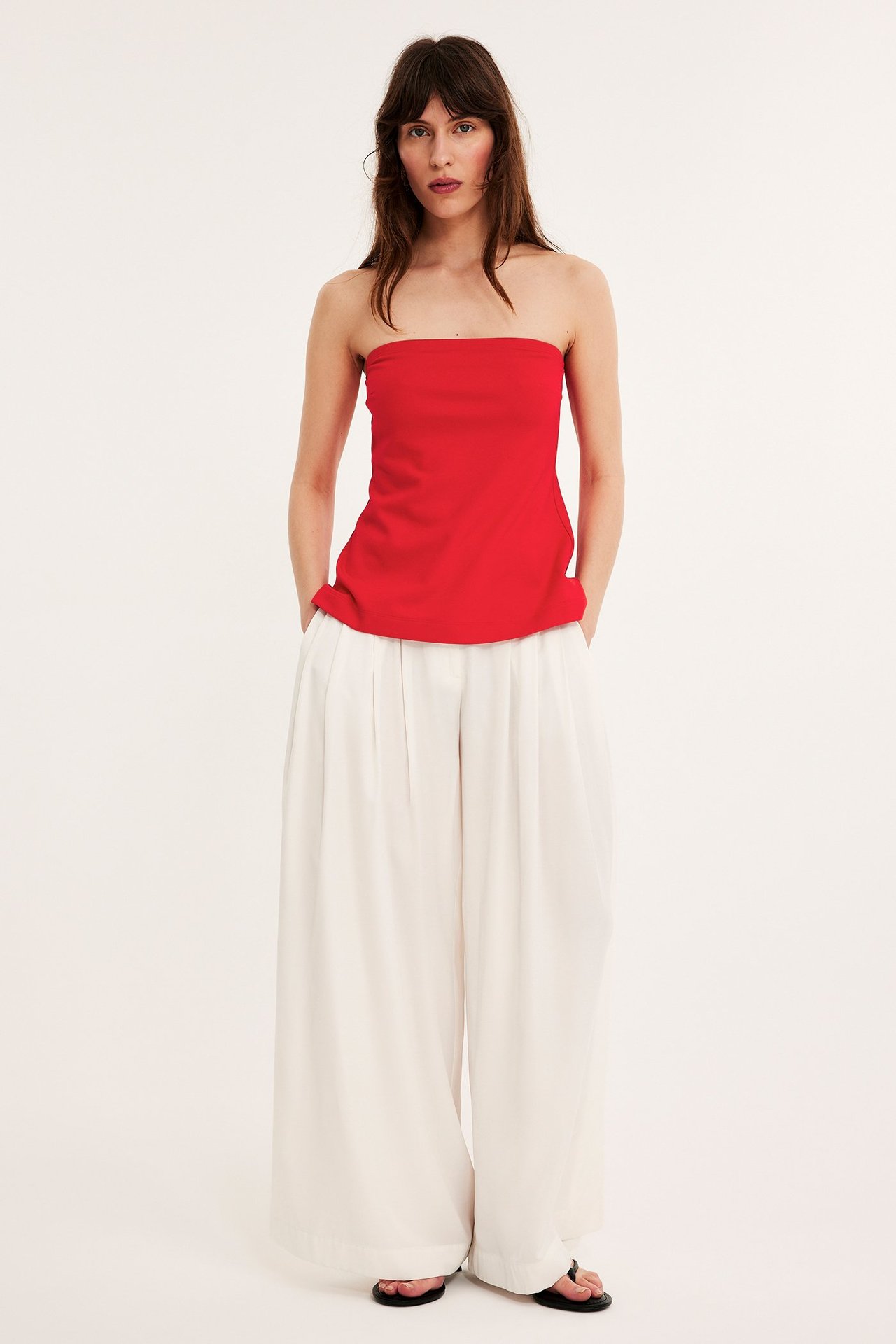 Widok z przodu: kobieta w czerwonym dłuższym topie bez ramiączek, białych szerokich spodniach i czarnych klapkach.