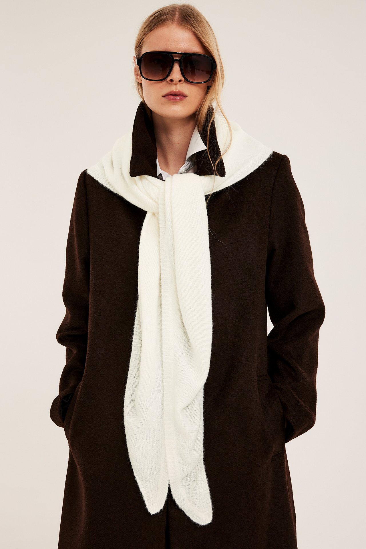 Nainen edestä: Yksivärinen luonnonvalkoinen kolmiohuivi valkoisen paidan ja tummanruskean takin päällä, aurinkolasit.