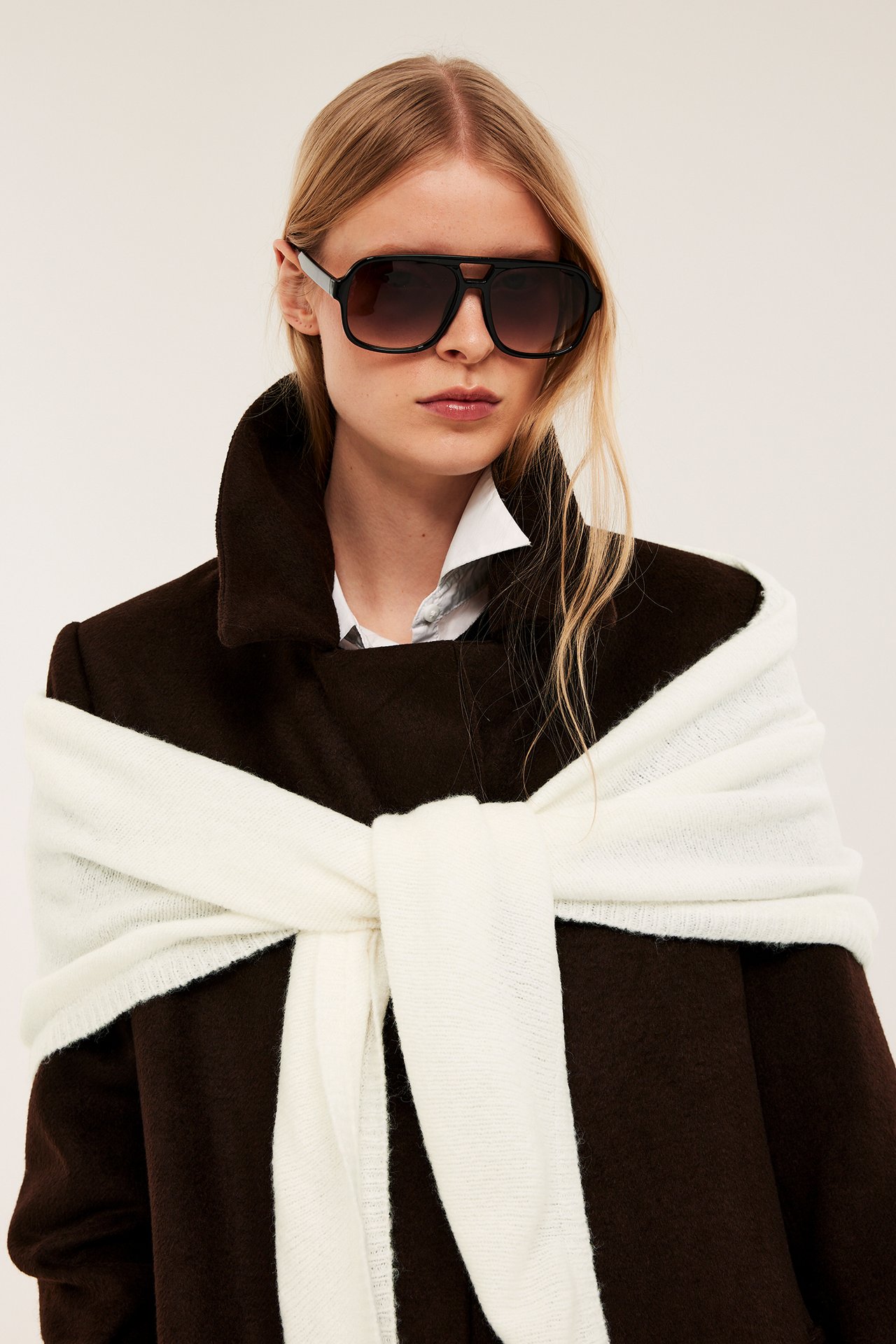 Frontvy: kvinna bär en offwhite enfärgad triangelscarf knuten över en brun kappa, med solglasögon.