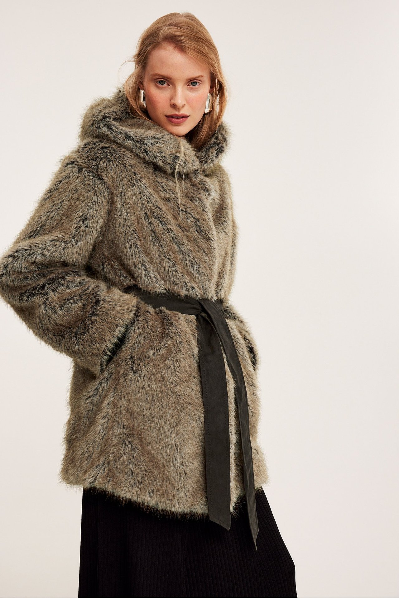 Edestä: Nainen beigessä tekoturkishupullisessa takissa mustalla solmimisvyöllä.