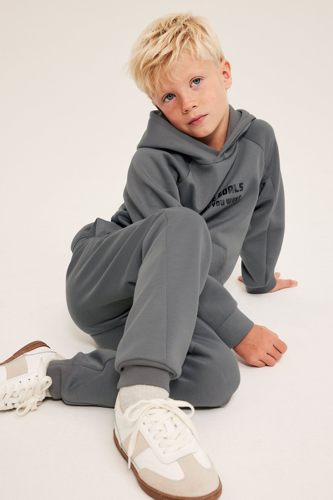 Istuva lapsi harmaassa interlock-hupparissa printillä, yhteensopivissa housuissa ja valkoisissa tennareissa.