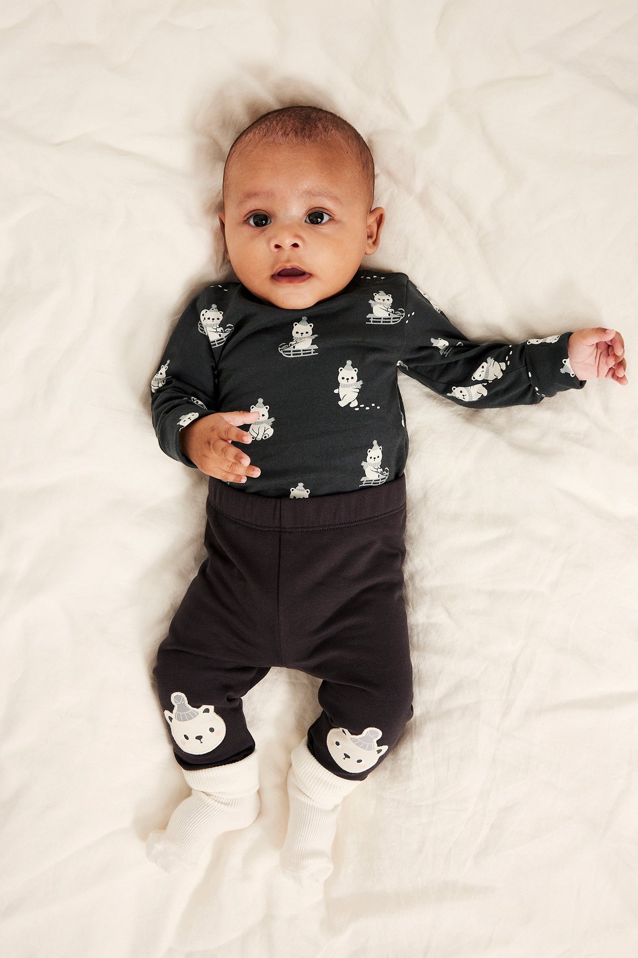 Framsida: baby i mörkgrå långärmad body med vitt nallebjörnsmönster, mörkgrå byxor och vita strumpor.