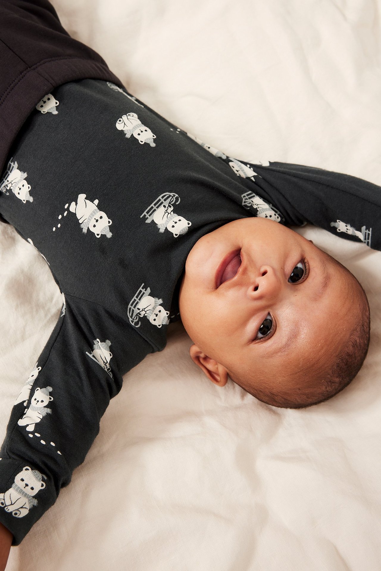Uppifrån, mörkgrå långärmad babybody med vitt nallebjörnsmönster på baby med mörka byxor.