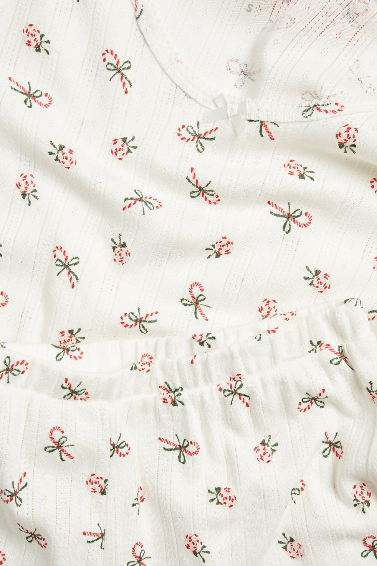 Lähikuva valkoisesta pointelle-neulepyjamasta, jossa punavihreitä karkkikeppi- ja rusettikuvioita sekä röyhelöitä.
