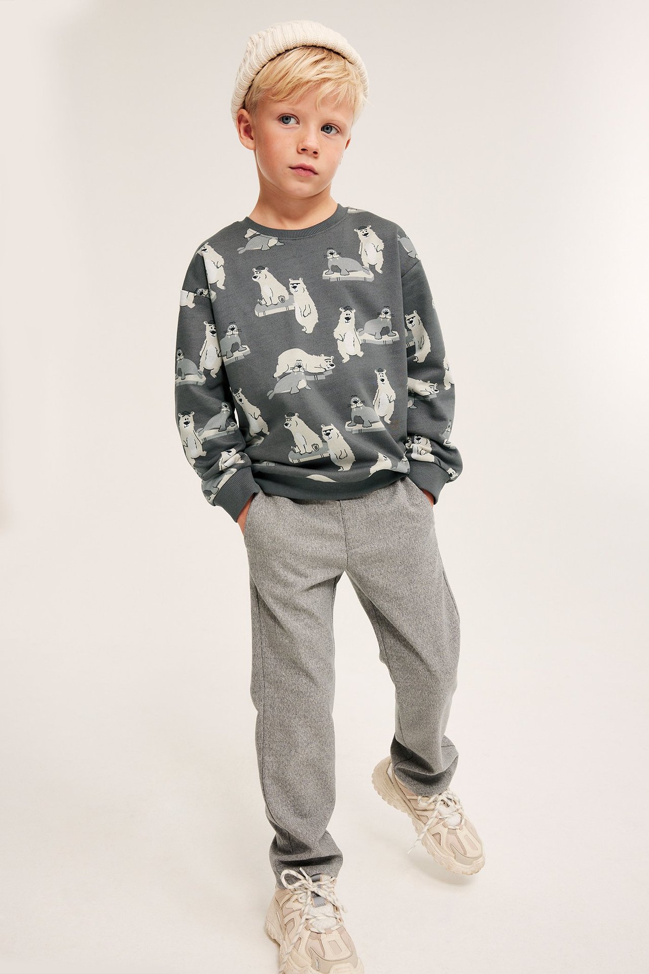 Widok z przodu: Dziecko w szarej bluzie z niedźwiedziami polarnymi, szarych spodniach, kremowej czapce, beżowych sneakersach.