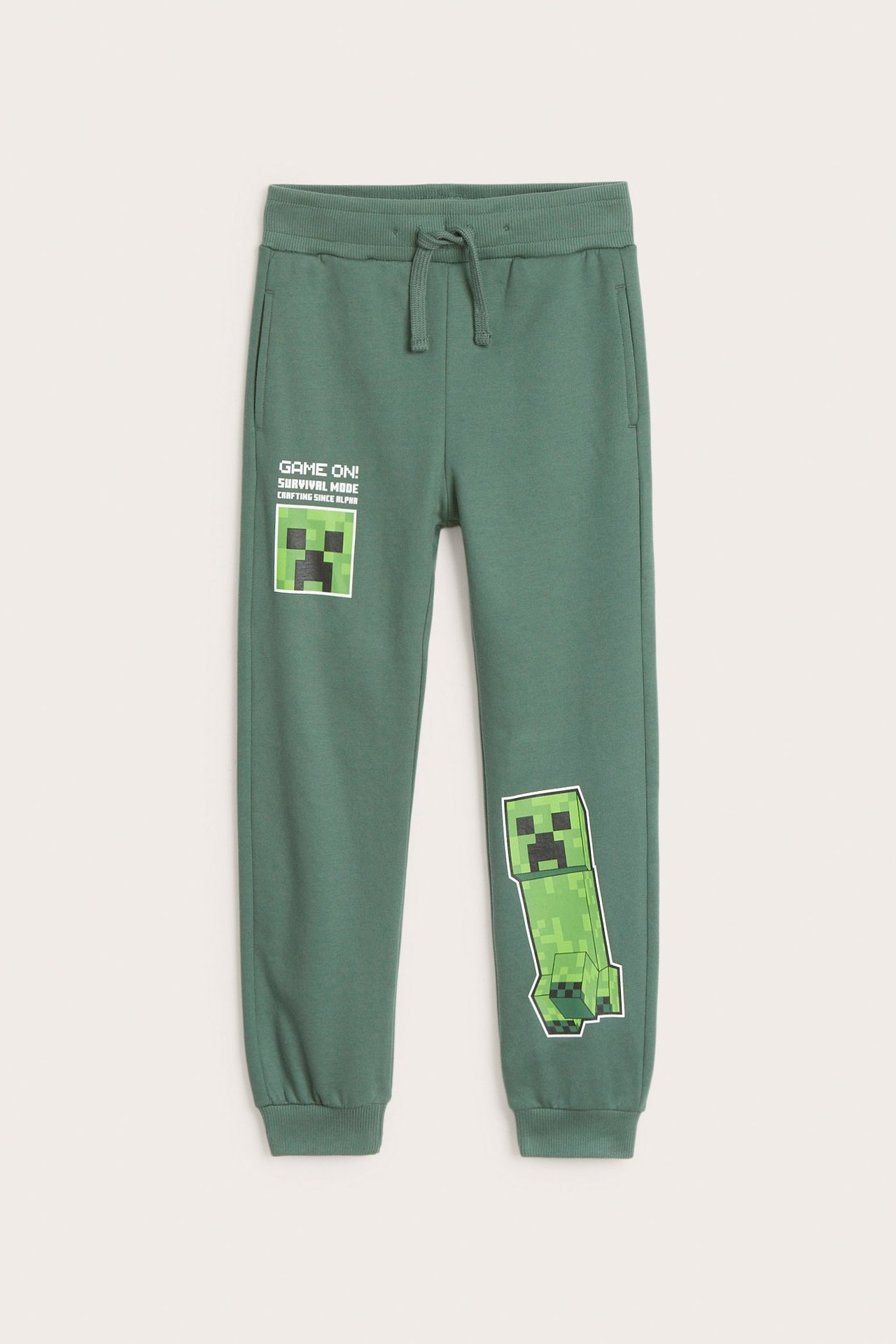 Edestä: tummanvihreät lasten puuvillaiset collegehousut Minecraft Creeper -painatuksella ja kiristysnyörillä vyötäröllä.