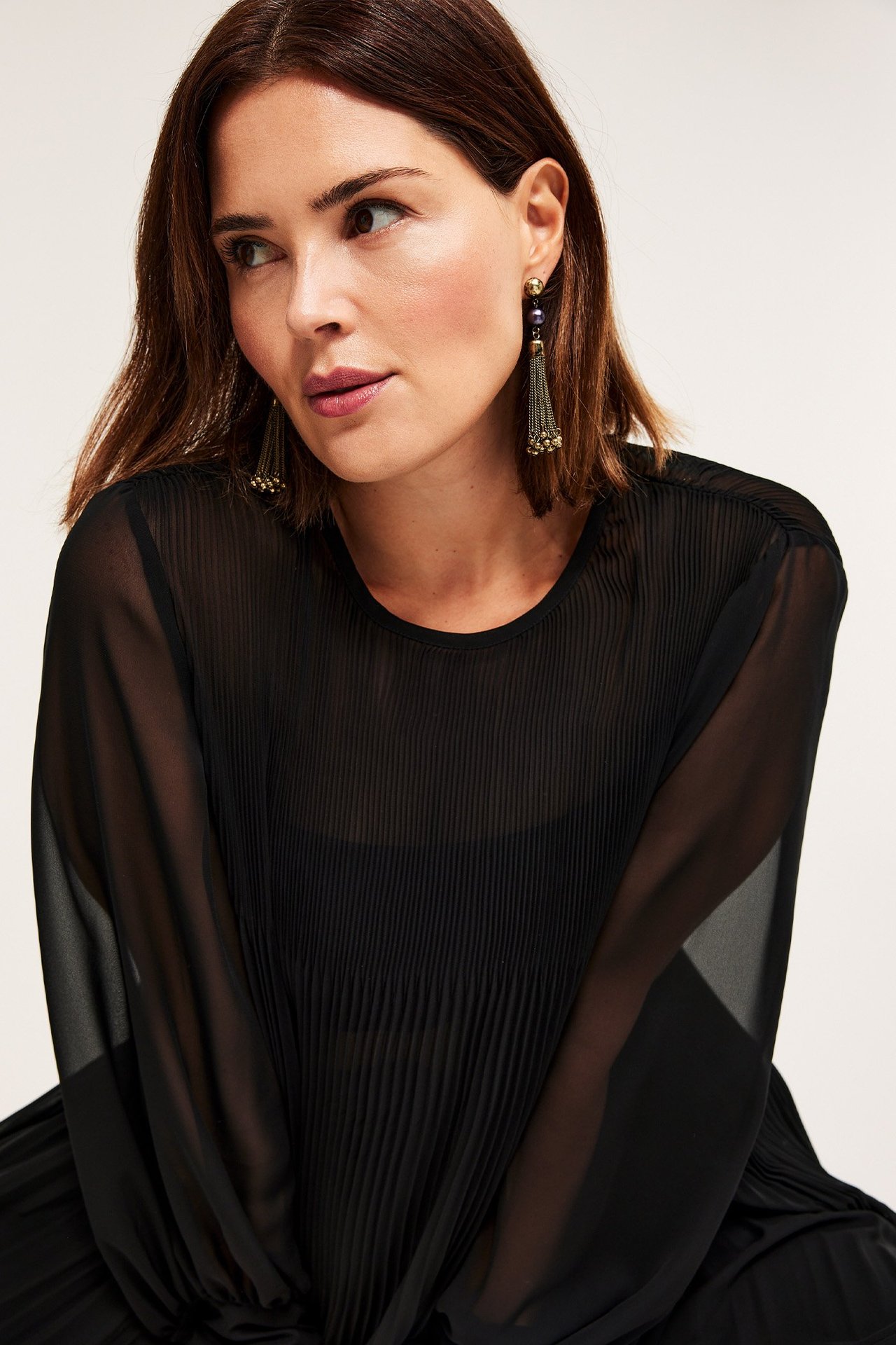 Lähikuva naisesta pitkissä kullanvärisissä tupsukorvakoruissa ja mustassa läpinäkyvässä laskostopissa.