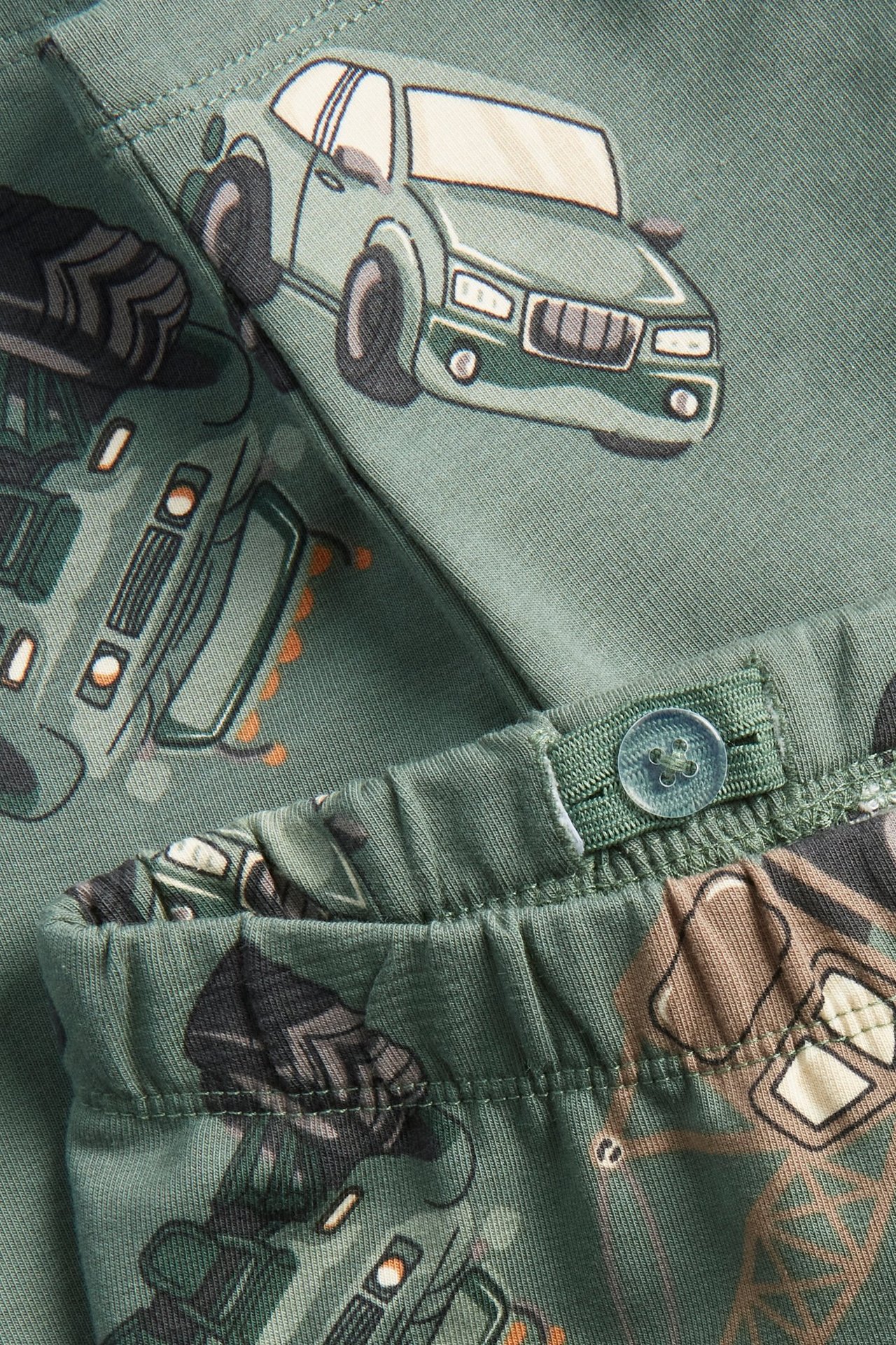 Zbliżenie na ciemnozielone legginsy dziecięce z nadrukiem samochodów i pojazdów budowlanych, regulowany pas z guzikiem.