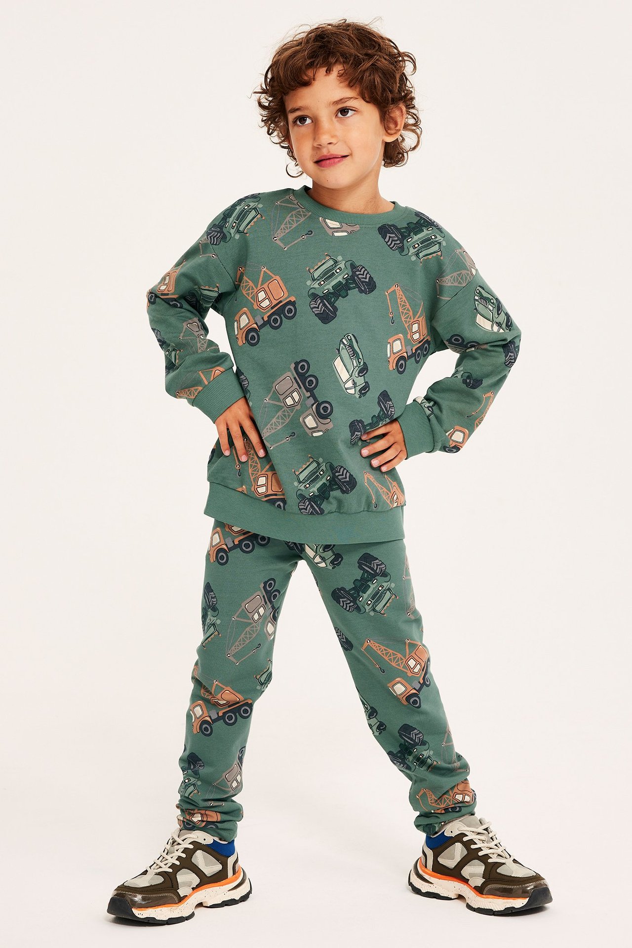 Widok z przodu: Dziecko w ciemnozielonej bluzie i pasujących legginsach we wzór pojazdów budowlanych, oraz wielokolorowe sneakersy.