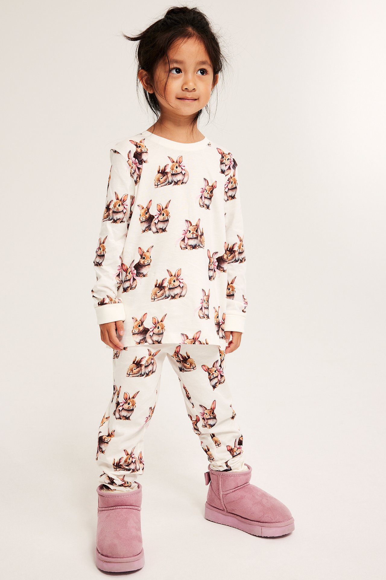Framifrån, ett barn i offwhite långärmad pyjamas i bomull med kanintryck och rosa stövlar.