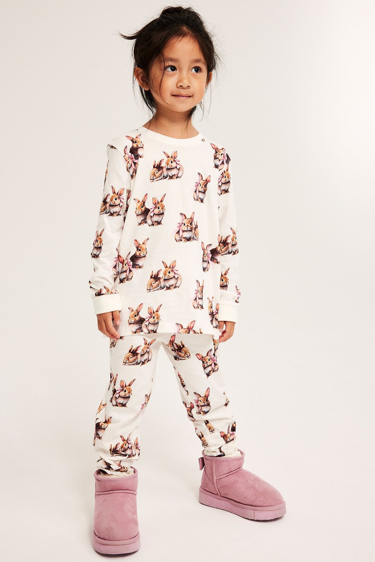 Edestä, lapsi yllään luonnonvalkoinen pitkähihainen puuvillapyjama pupupainatuksella ja vaaleanpunaiset saappaat.