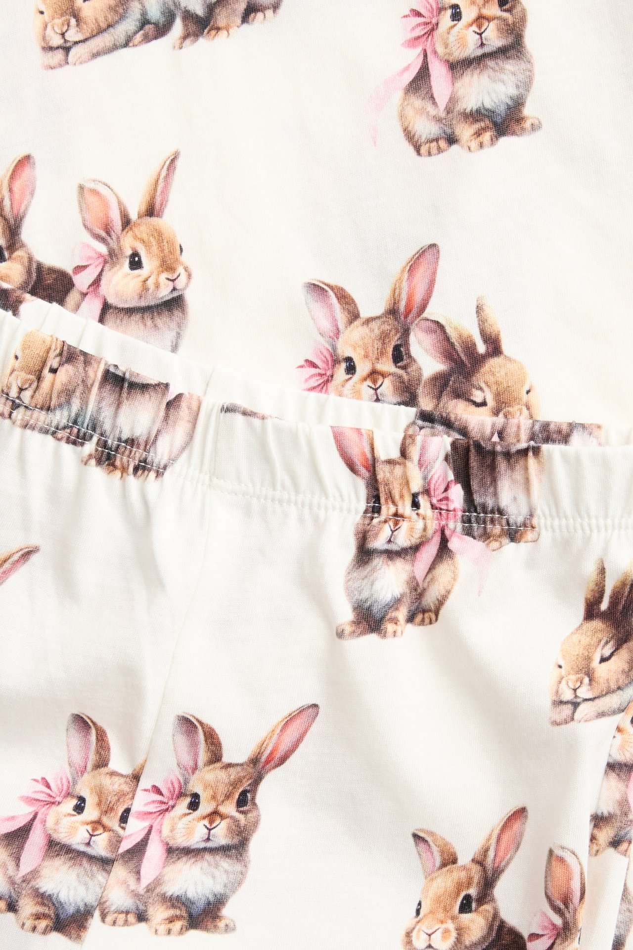 Zbliżenie na ecru bawełnianą piżamę dziecięcą z powtarzającym się wzorem brązowych królików z różowymi kokardami.