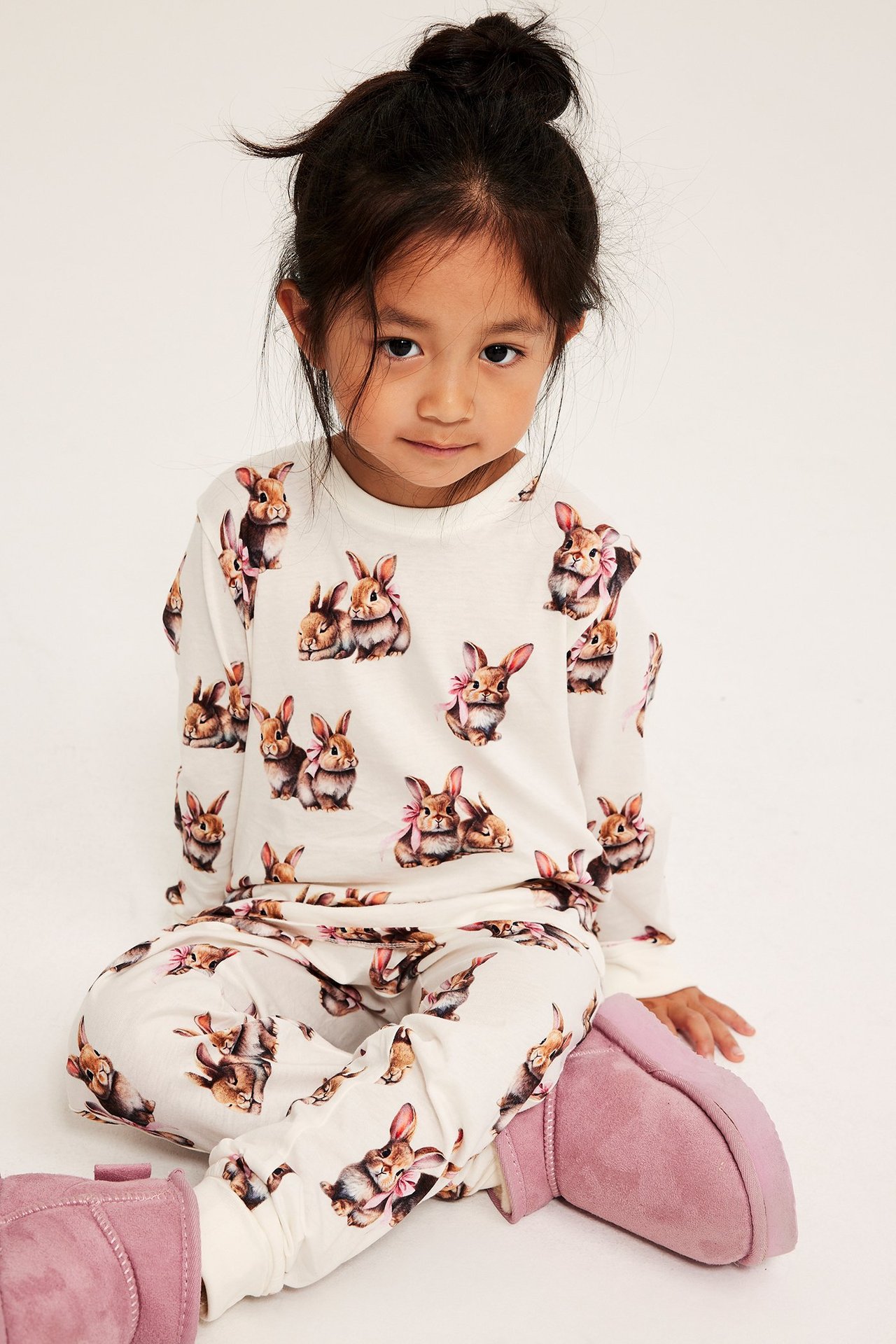 Framifrån, ett barn i offwhite pyjamas av bomull med kaninmönster och rosa stövlar.