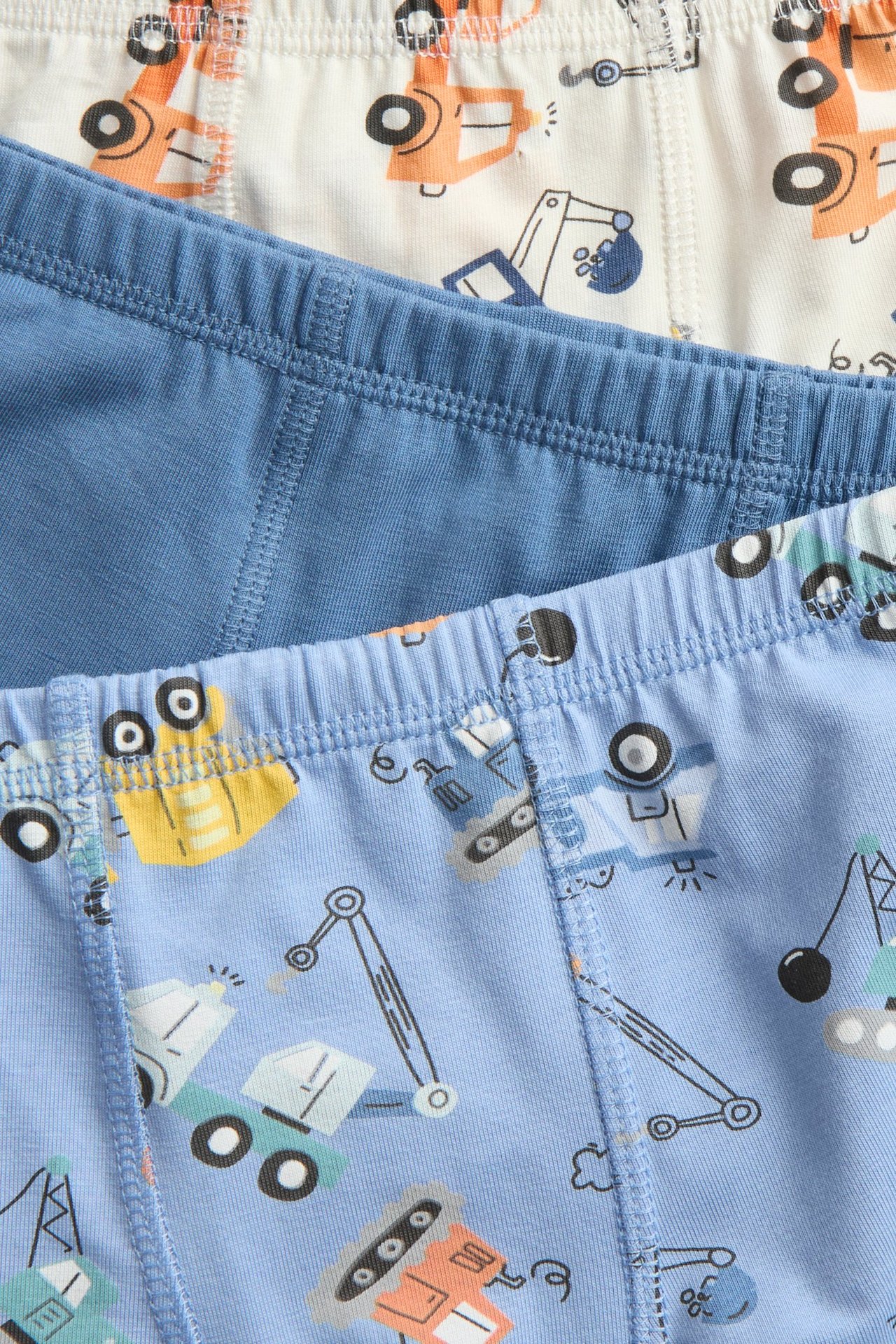 Närbild på tre par barnboxerkalsonger: ljusblå med fordonsmönster, enfärgad blå och benvit med orange fordonsmönster.