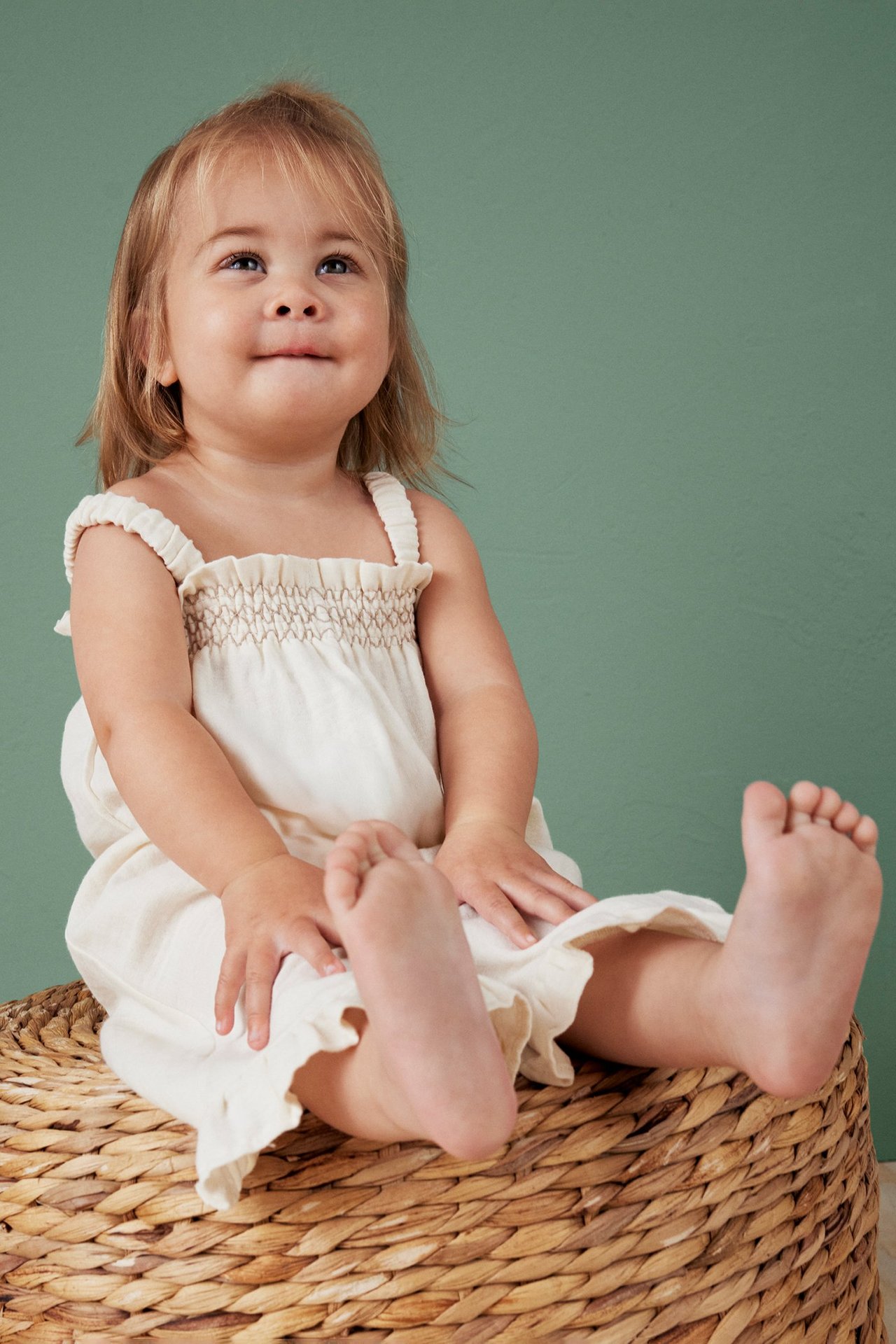 Edestä, vauva yllään beige puuvillakreppinen haalari röyhelöolkaimilla ja smokkiryppytyksellä, istuen korissa.