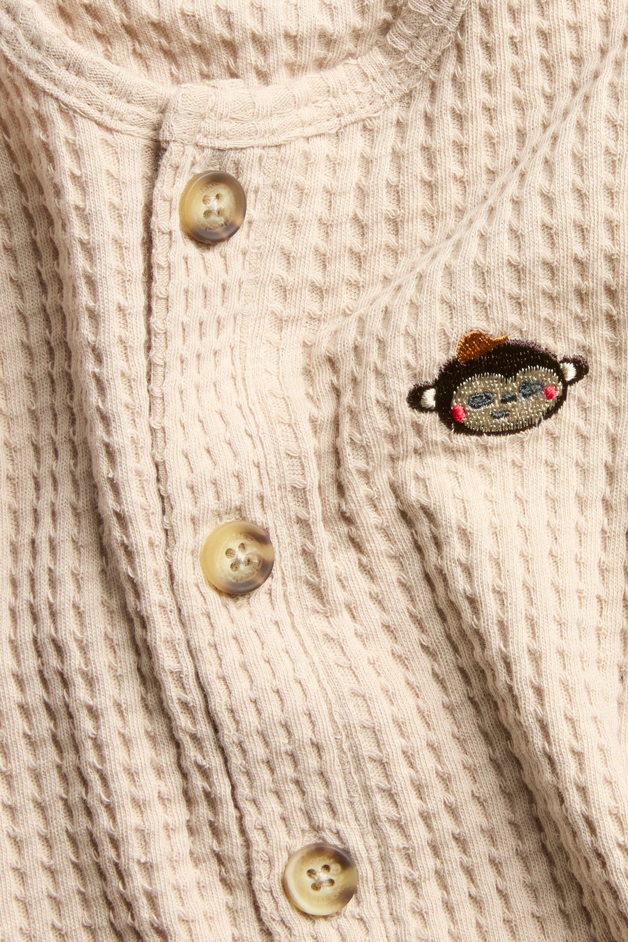 Lähikuva beigestä vohvelikankaisesta vauvan haalarista, jossa edessä napit ja apinakirjonta rinnassa.